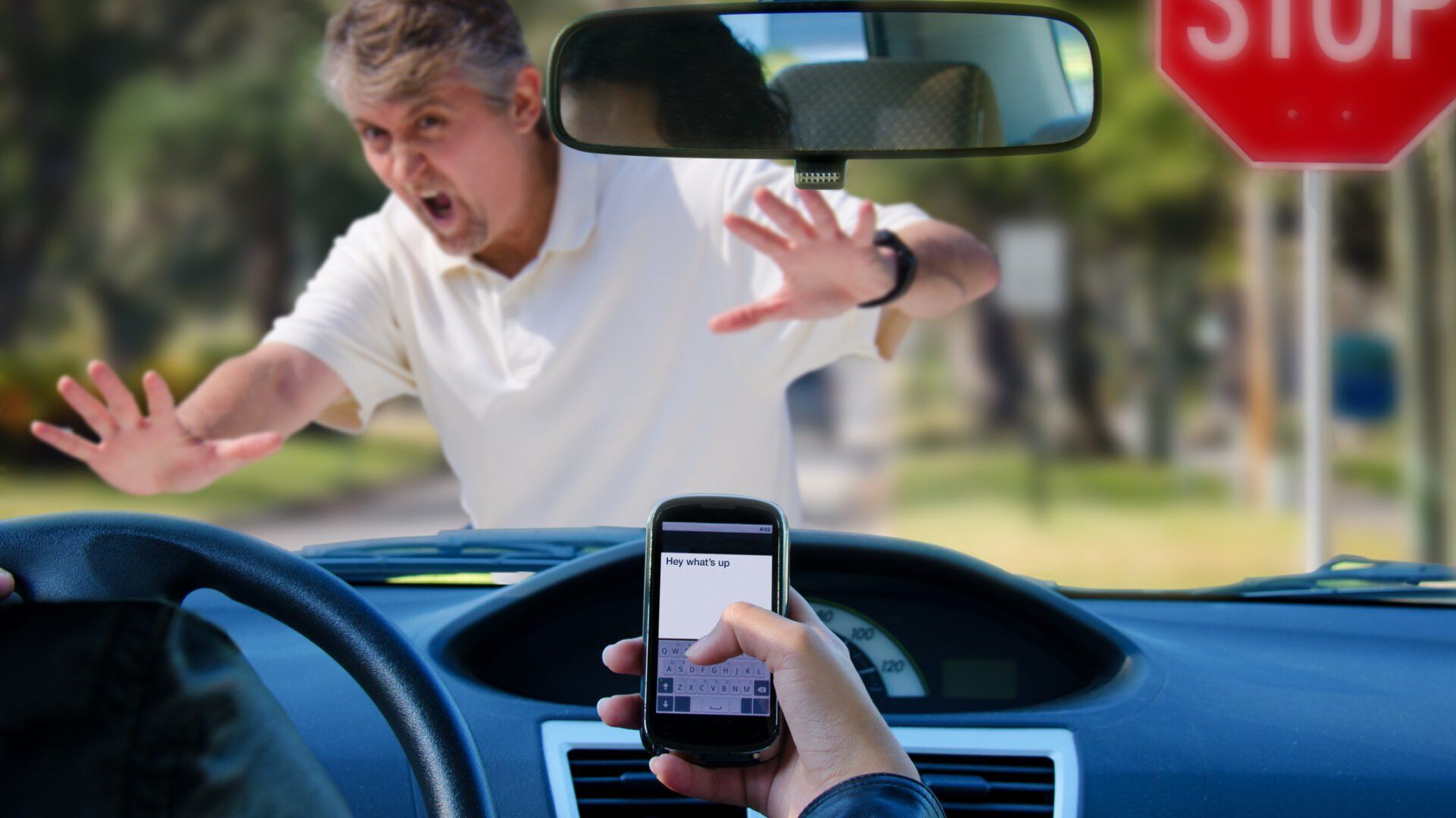 A driver looks at a phone while a person stands in front of the car with hands raised in alarm near a stop sign.