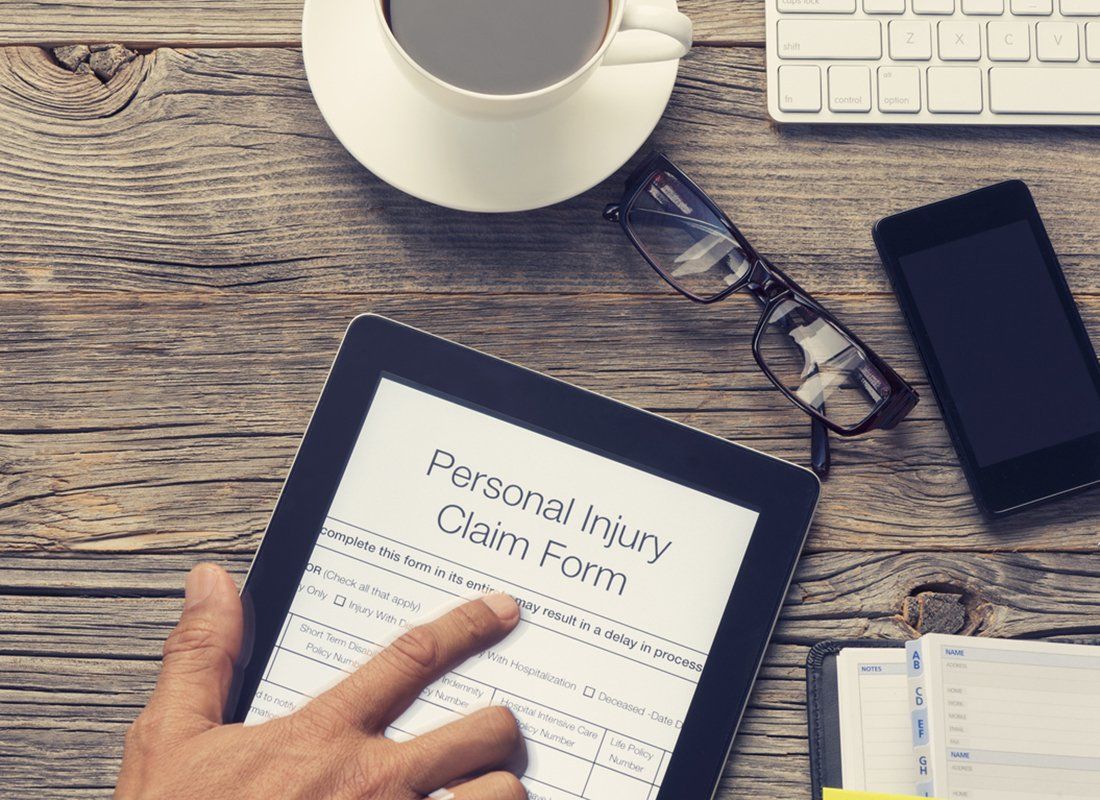 A hand uses a digital tablet to fill out a Personal Injury Claim Form on a wooden desk with a coffee mug and glasses.