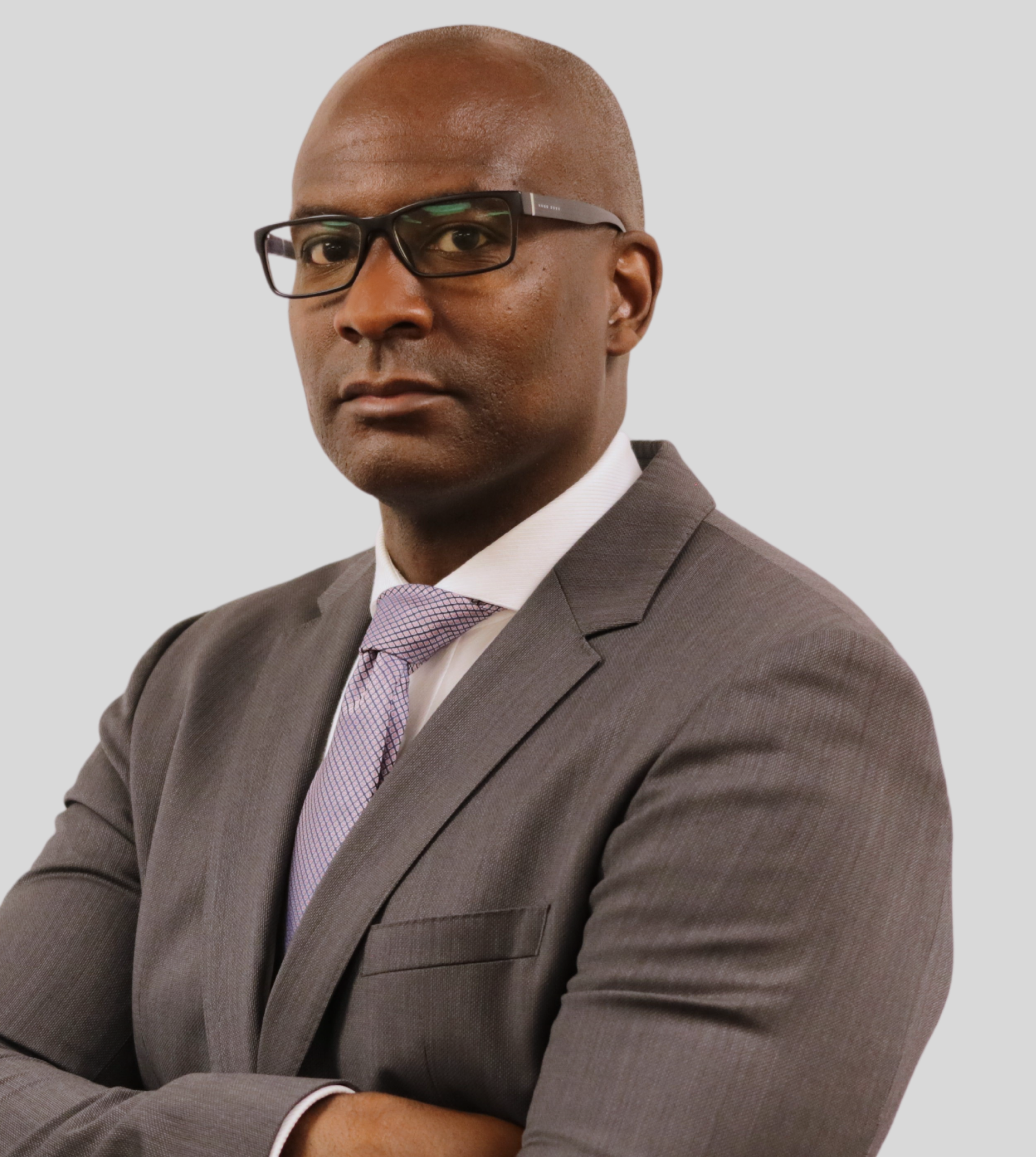 A person in a gray suit and patterned tie with glasses, arms crossed, looking toward the camera against a gray background.