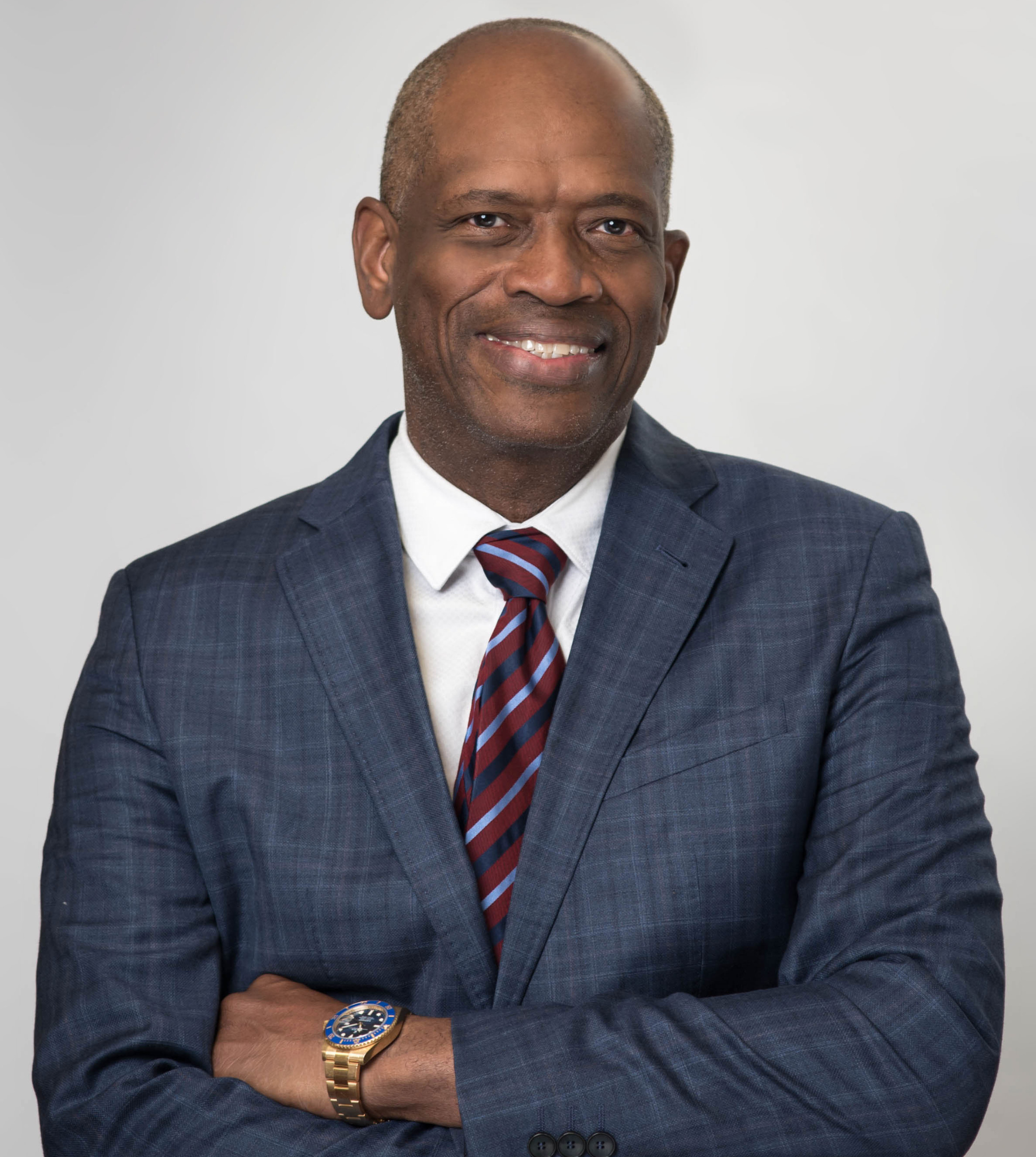 A smiling man in a blue textured blazer, white shirt, and striped tie with arms crossed, set against a light background.