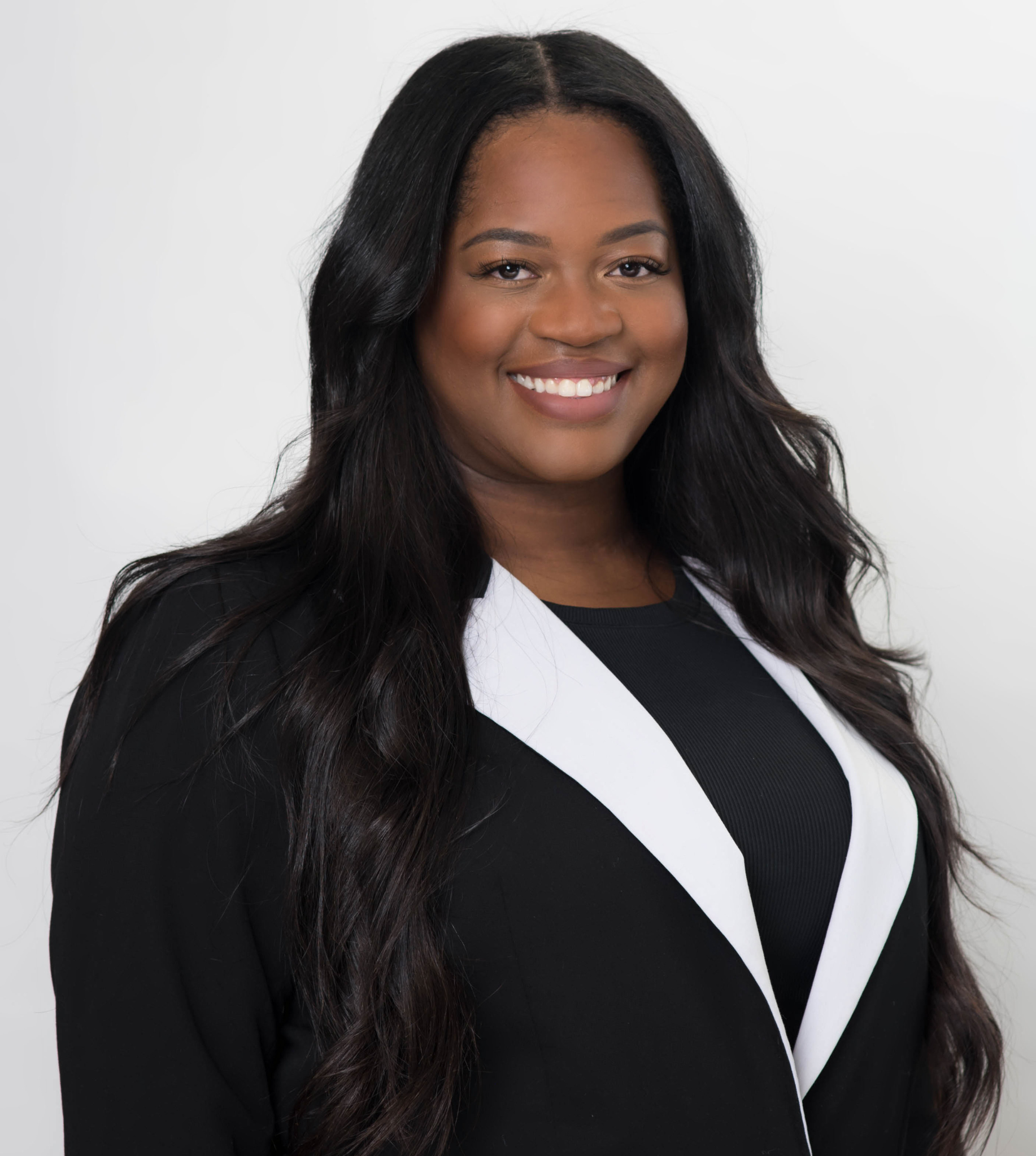 A person with long, dark hair wearing a black blazer with white lapels, smiling against a plain white background.