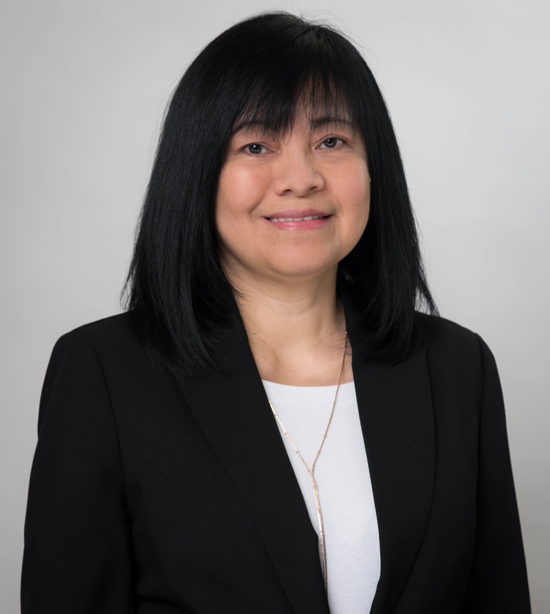 A smiling woman with dark hair, wearing a white shirt and black blazer against a light gray background.
