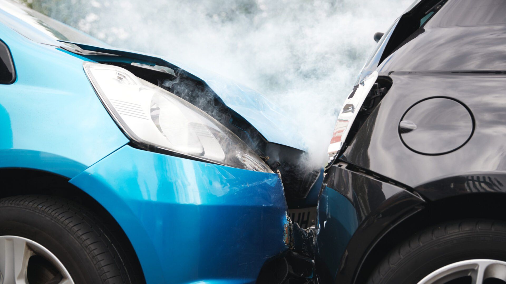 A close-up view of a blue car and a black car in a collision, with smoke rising from the point of impact.