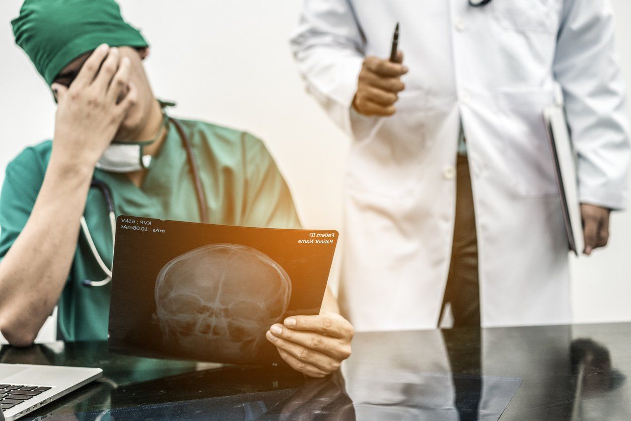 A medical professional in green scrubs covers their face in frustration while holding a skull X-ray near another doctor.