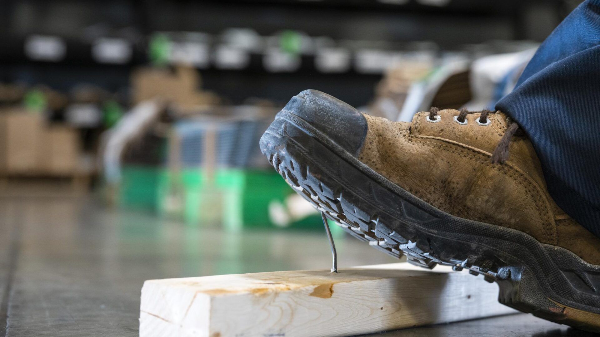 A tan work boot steps over a nail protruding from a piece of wood on a concrete floor in a warehouse setting.