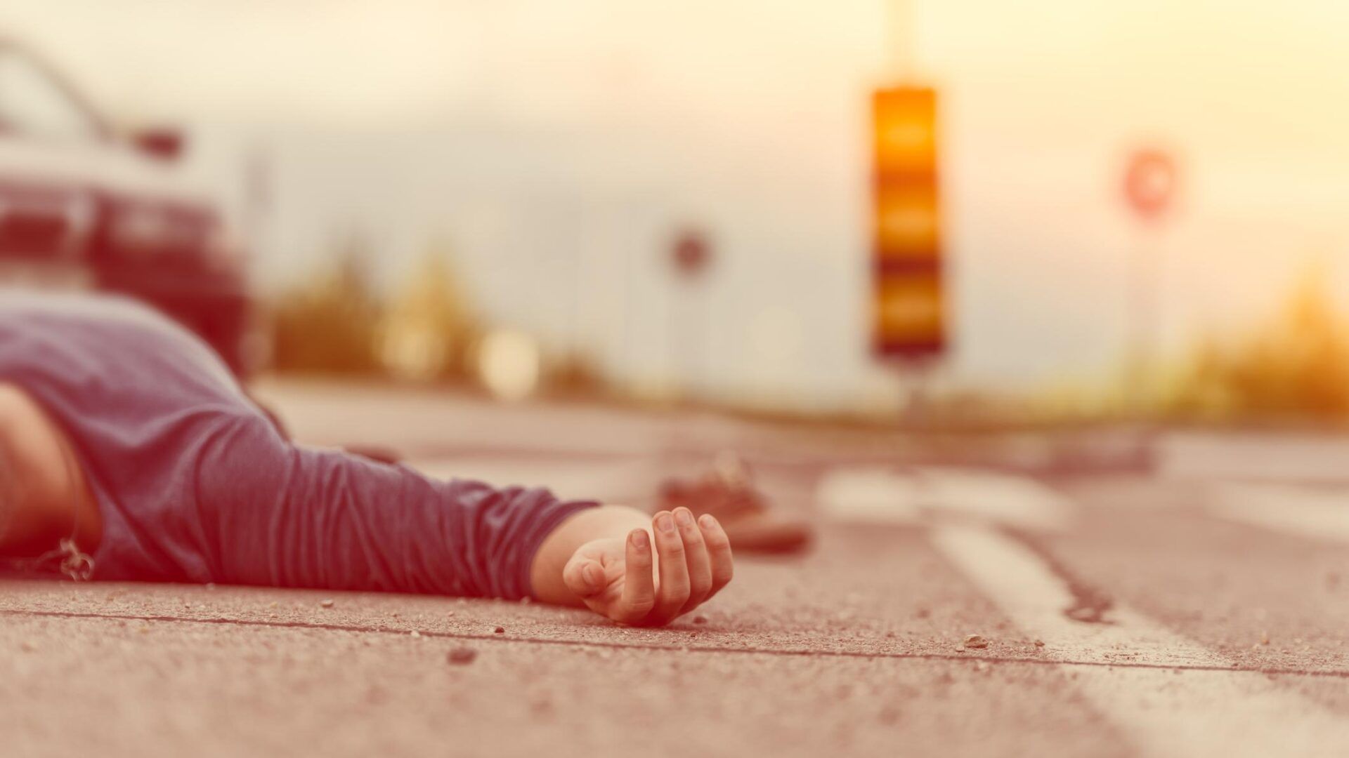 A person lies on the asphalt of a road near a crosswalk, with a blurred car and traffic light in the background.