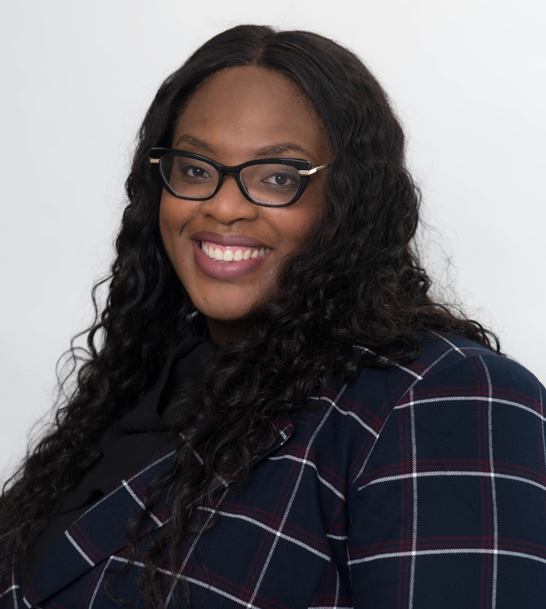 A smiling person with long dark hair and glasses wears a navy blue plaid blazer against a plain white background.