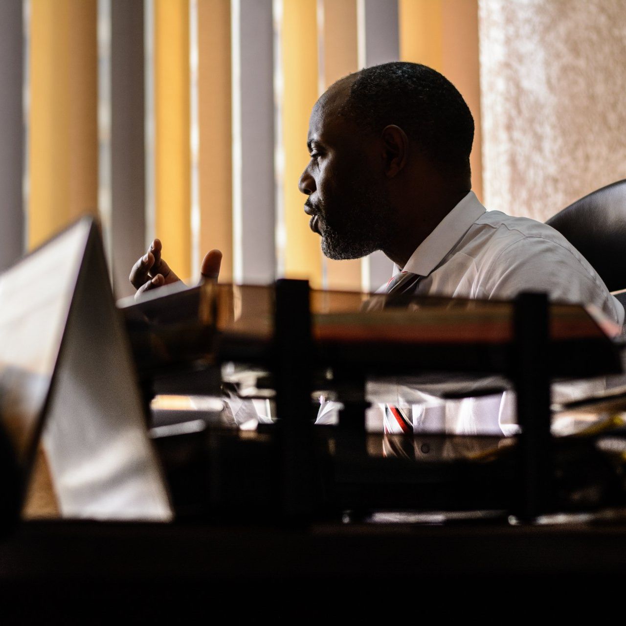A person in a white collared shirt sits at a desk, gesturing while looking toward a bright, slatted window.