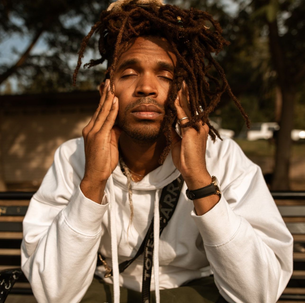 A person with long locs, eyes closed and hands touching their face, sits on a park bench wearing a white hoodie.