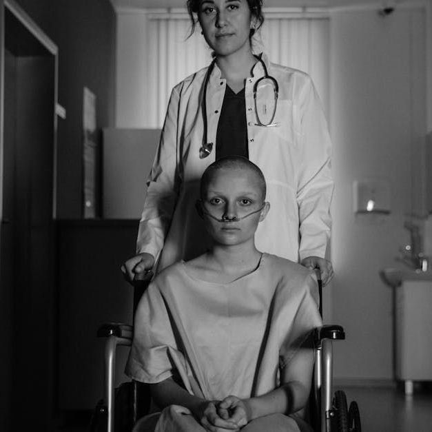 A patient with a shaved head and oxygen tube sits in a wheelchair, with a medical professional standing behind them.