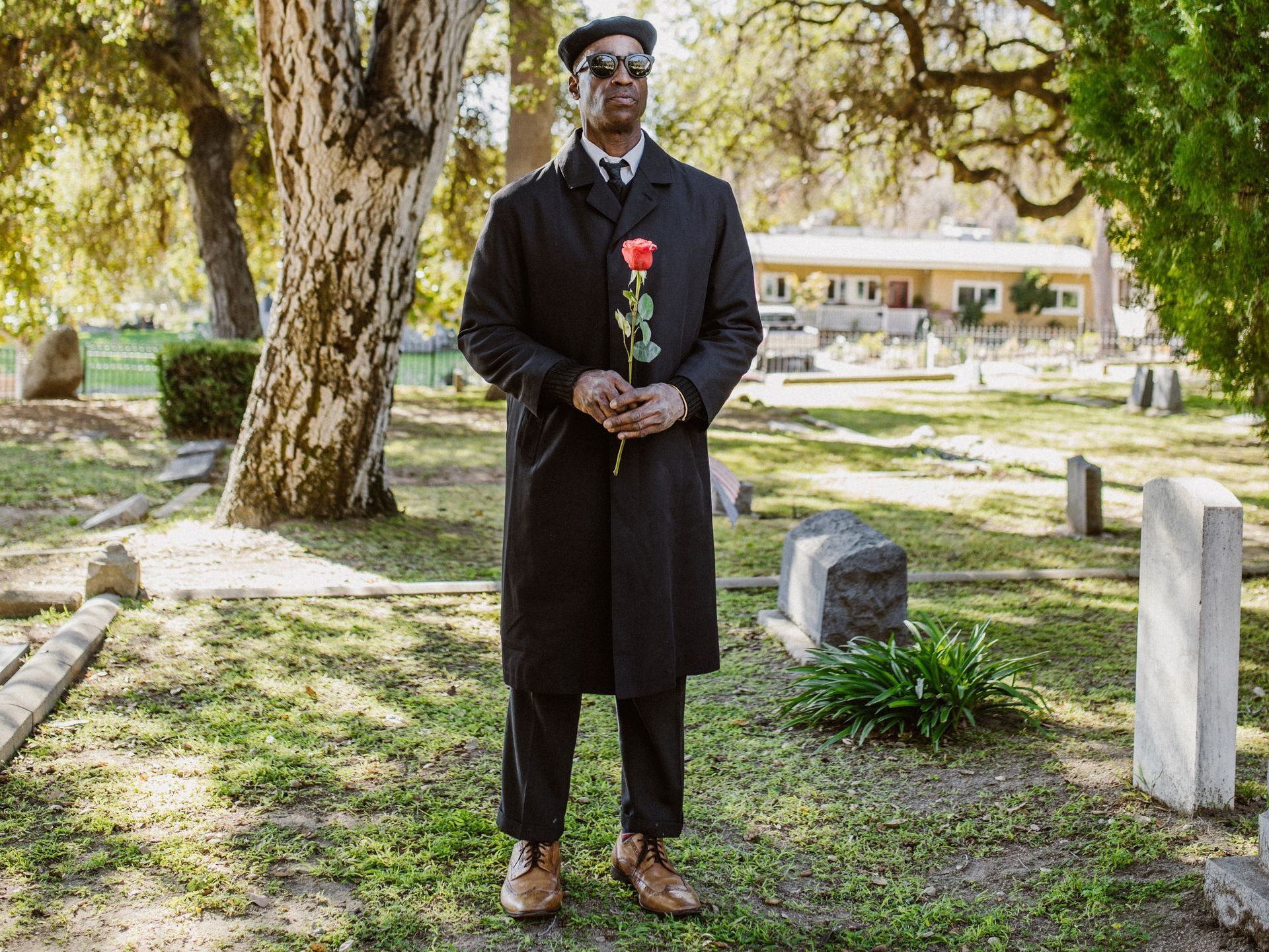 A person in a black coat and beret stands in a cemetery holding a single red rose.