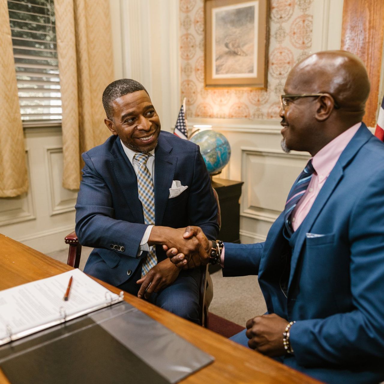 Two people in business suits shake hands across a desk in an office with a globe and framed artwork in the background.
