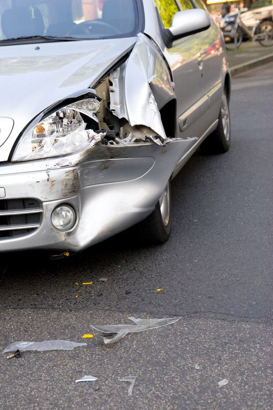 A silver car on a street with a severely damaged front fender, hood, and broken headlight.