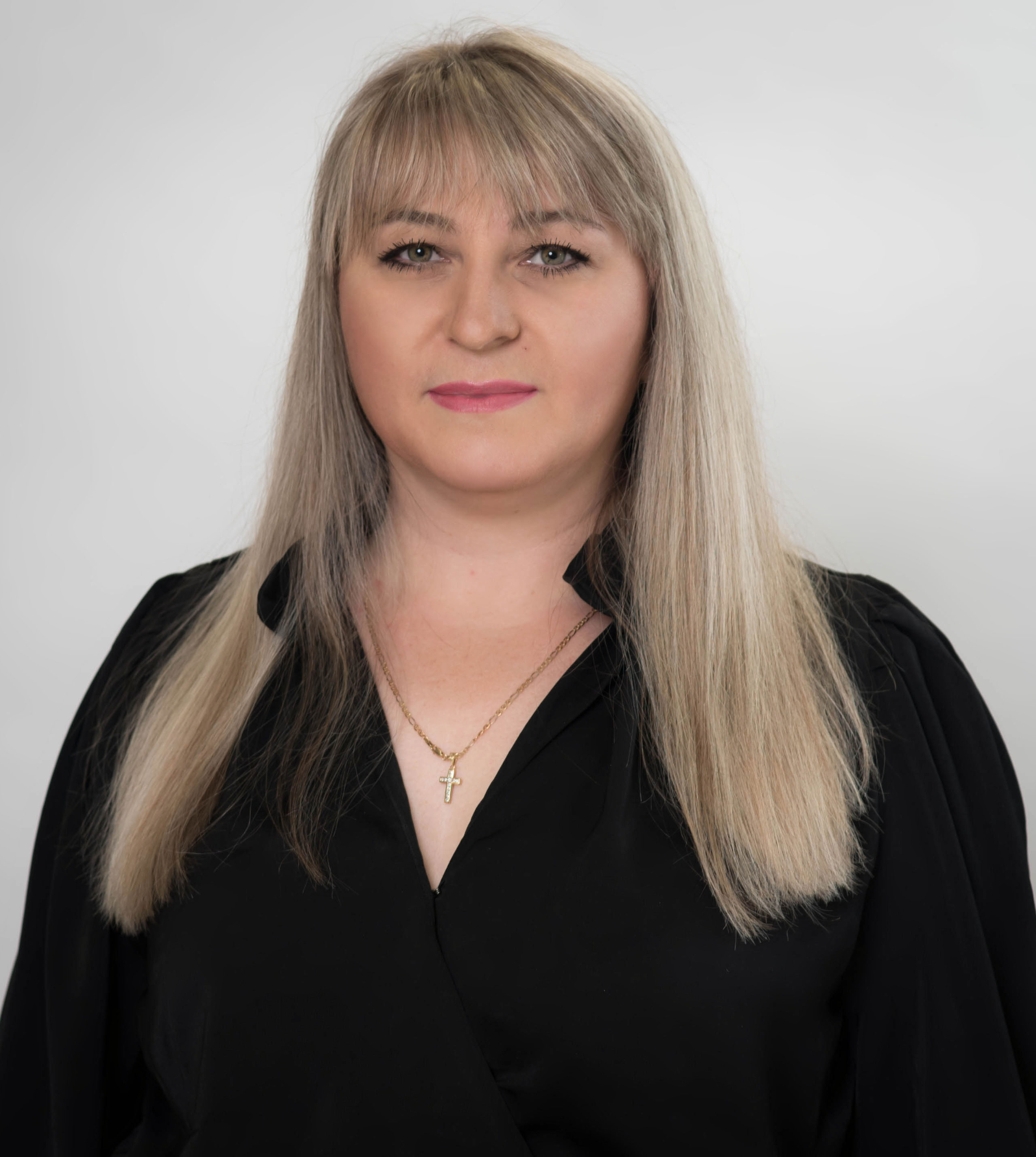 A person with shoulder-length blonde hair and bangs, wearing a black V-neck top and a delicate necklace against a gray wall.