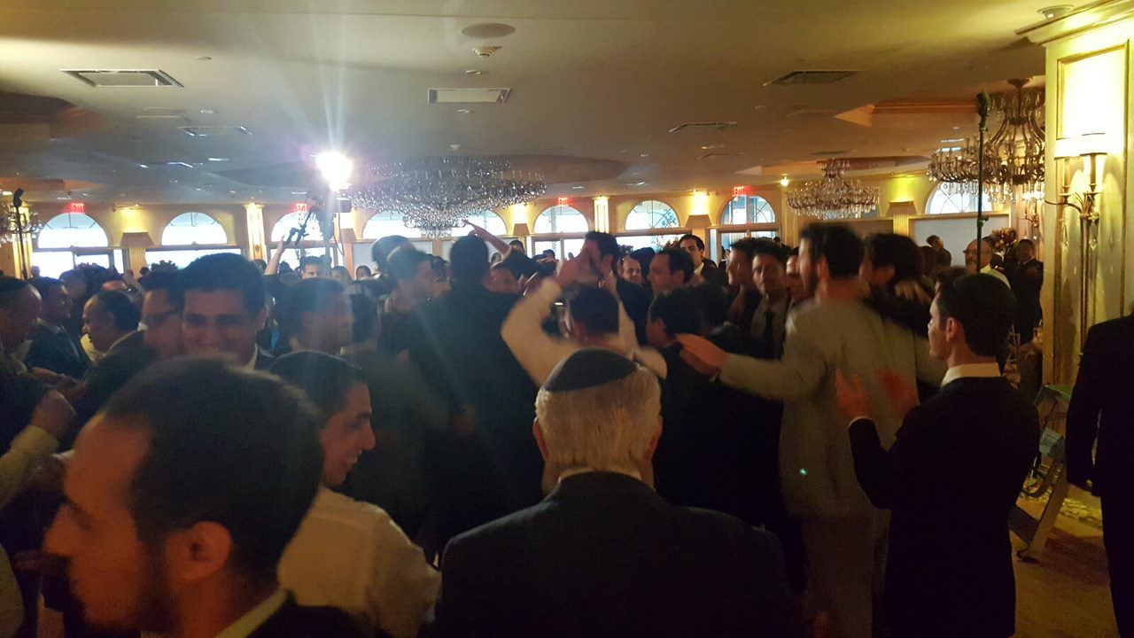 People dancing in a well-lit banquet hall with chandeliers and arched windows.