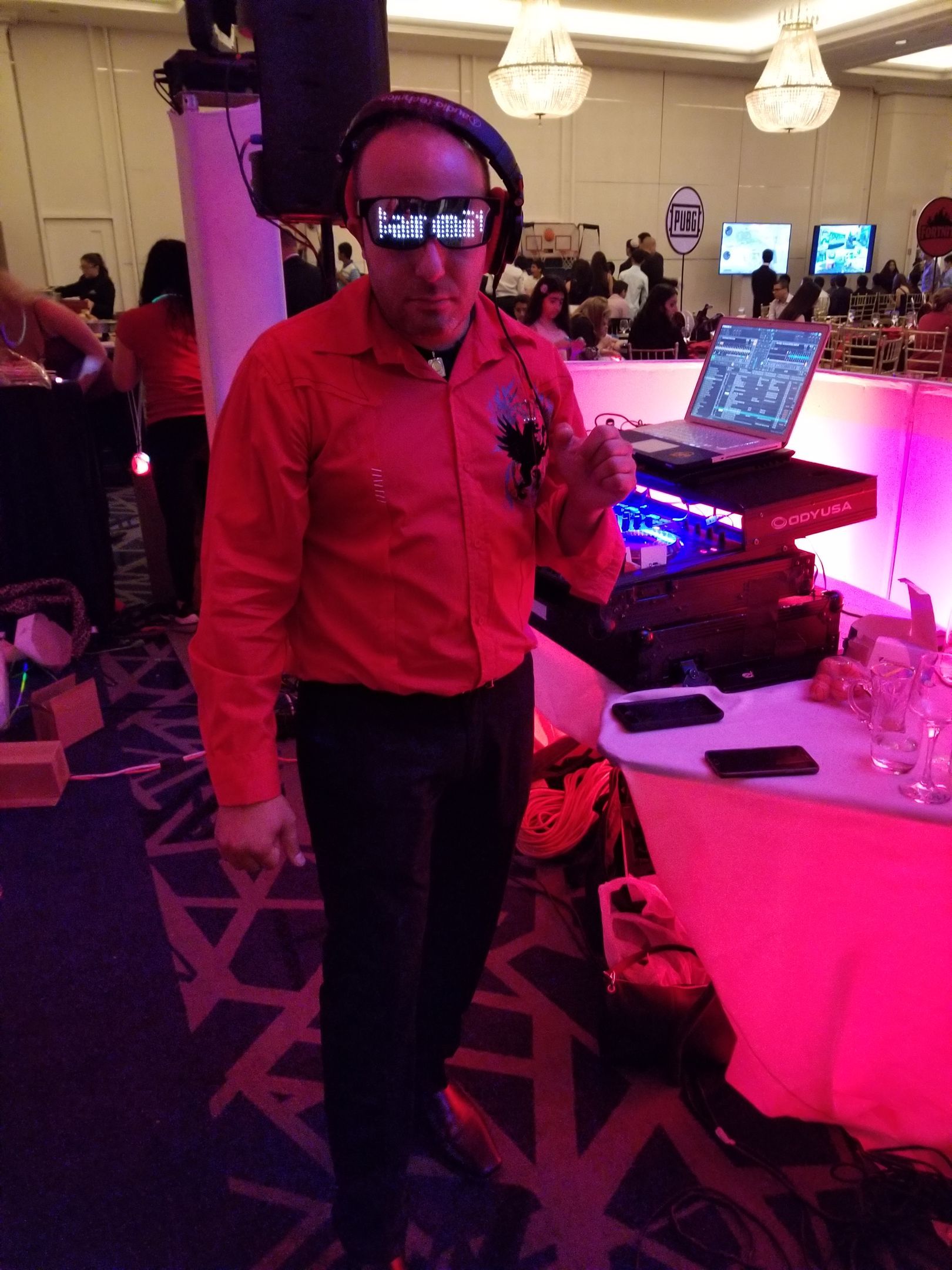 DJ wearing light-up glasses and headphones, in a red shirt, next to a laptop and DJ equipment.