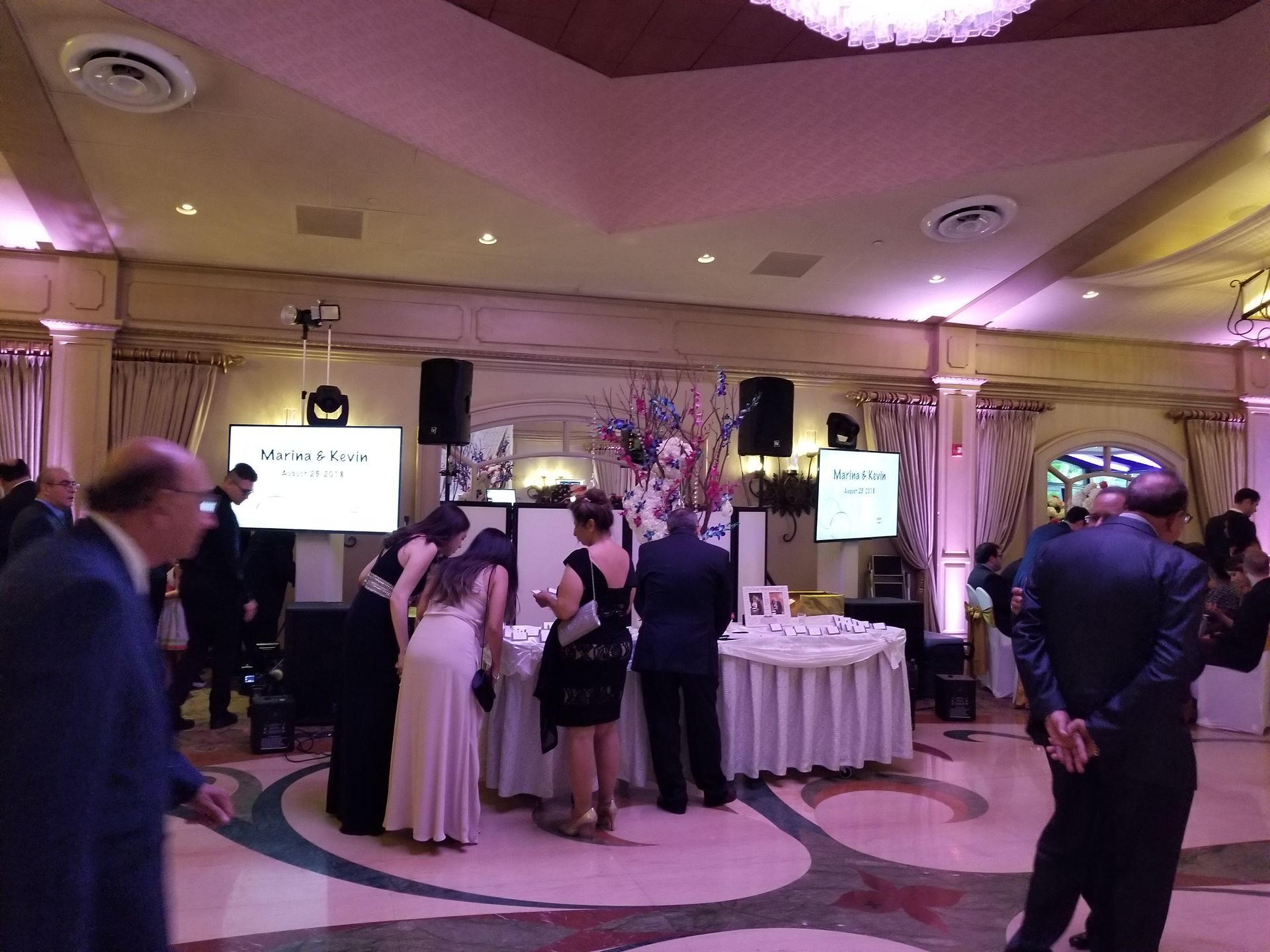 A banquet hall with people gathered around tables and screens, illuminated with pink lighting.