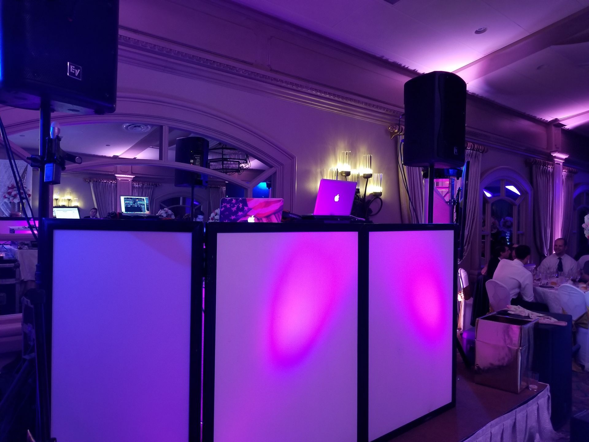 DJ setup at a party: white panels with purple uplighting, speakers, laptops, and a mirror in the background.