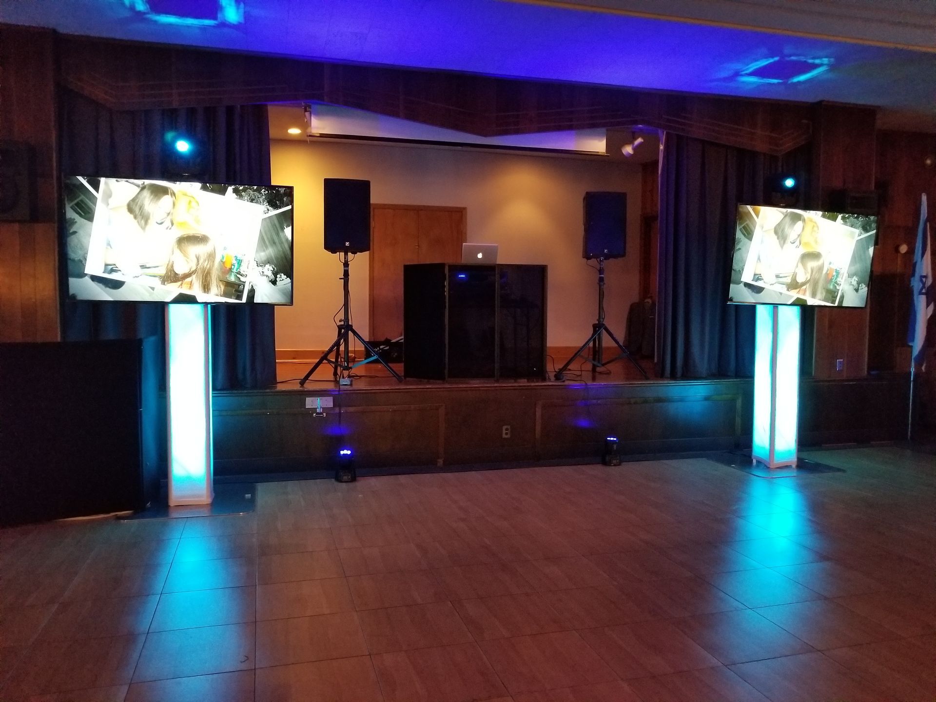 Stage setup with two screens displaying images, speakers, and DJ equipment illuminated with blue lights.
