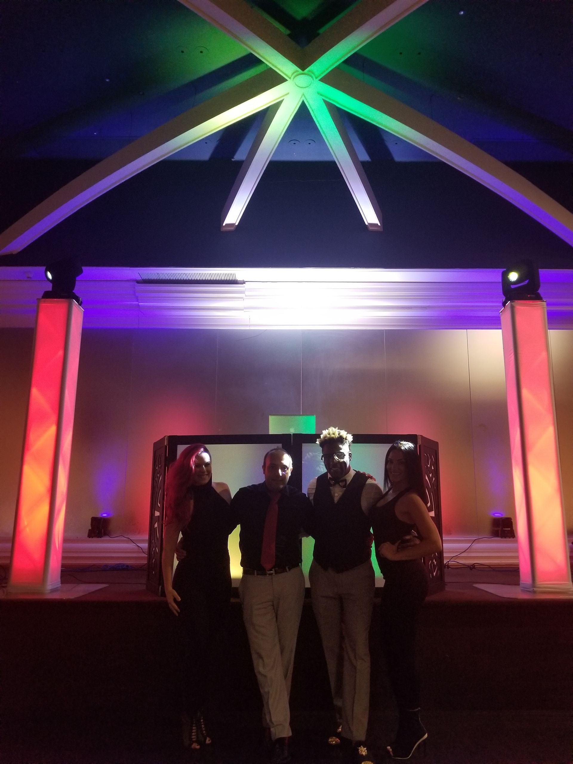 Four people standing on a stage, lit with colorful lights, arms around each other.