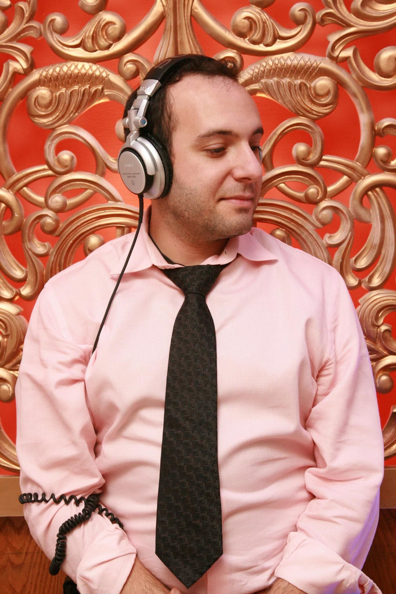 Man in pink shirt and tie wearing headphones, against a gold-colored ornate backdrop.
