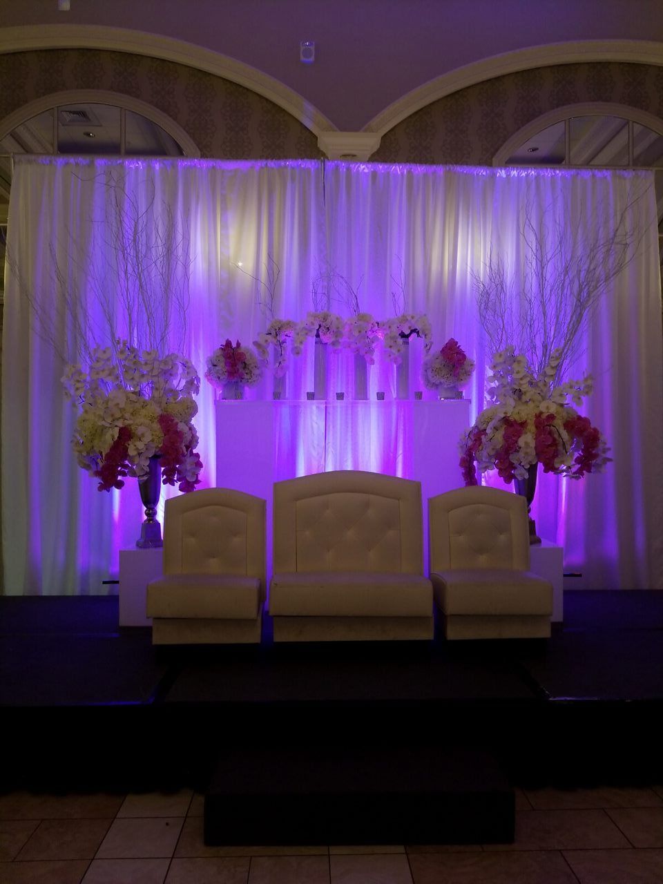 Wedding stage with white drapes, purple uplighting, chairs, and floral arrangements.