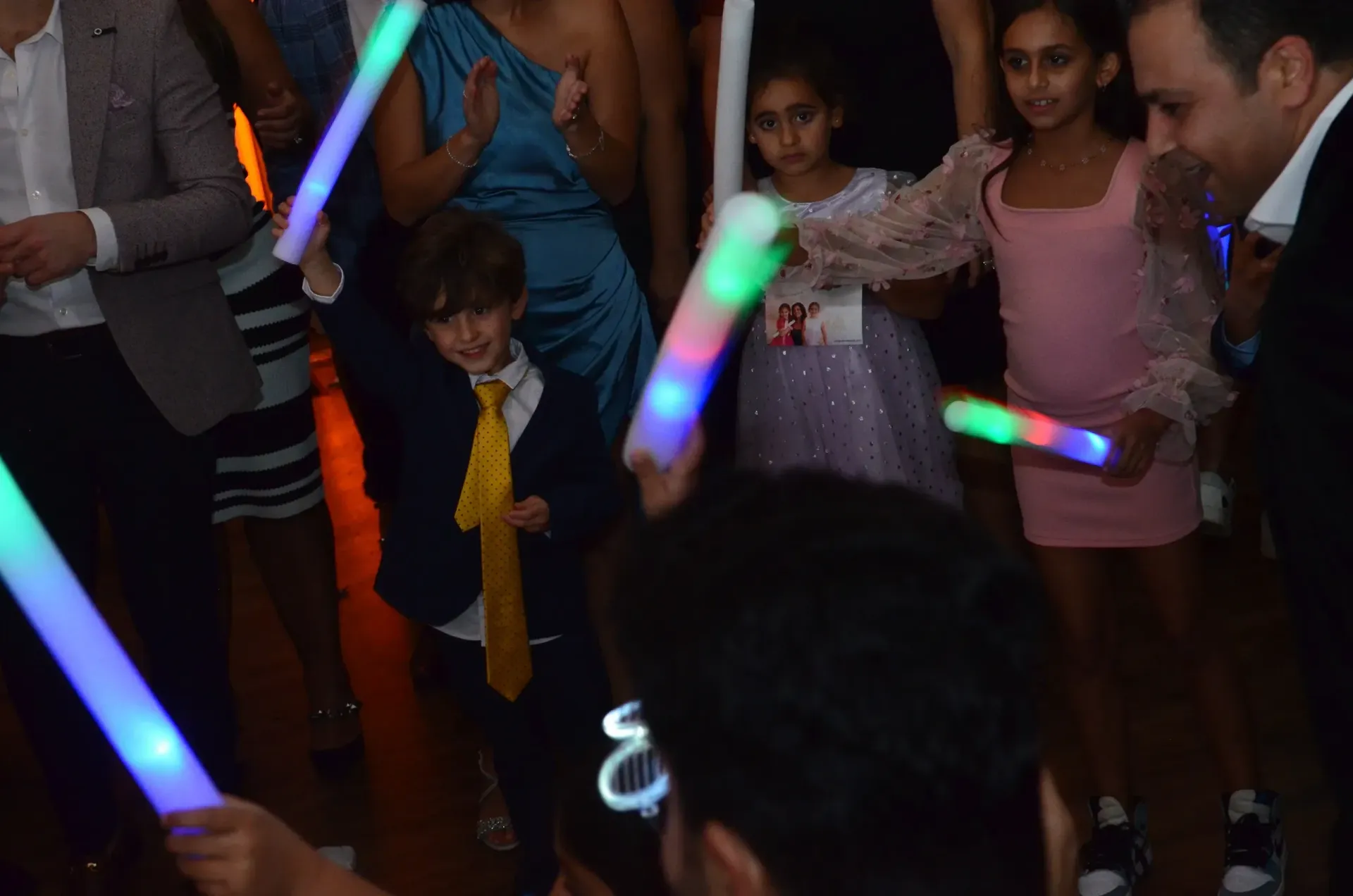People dancing at a party, holding glowing sticks. Boy in suit smiles. Others in dresses.