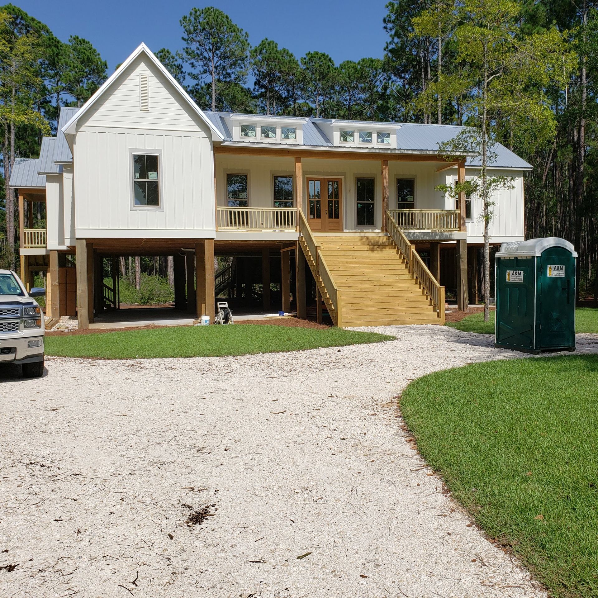 A newly constructed white elevated home with a wooden staircase, gravel driveway, and a portable toilet in the front yard.