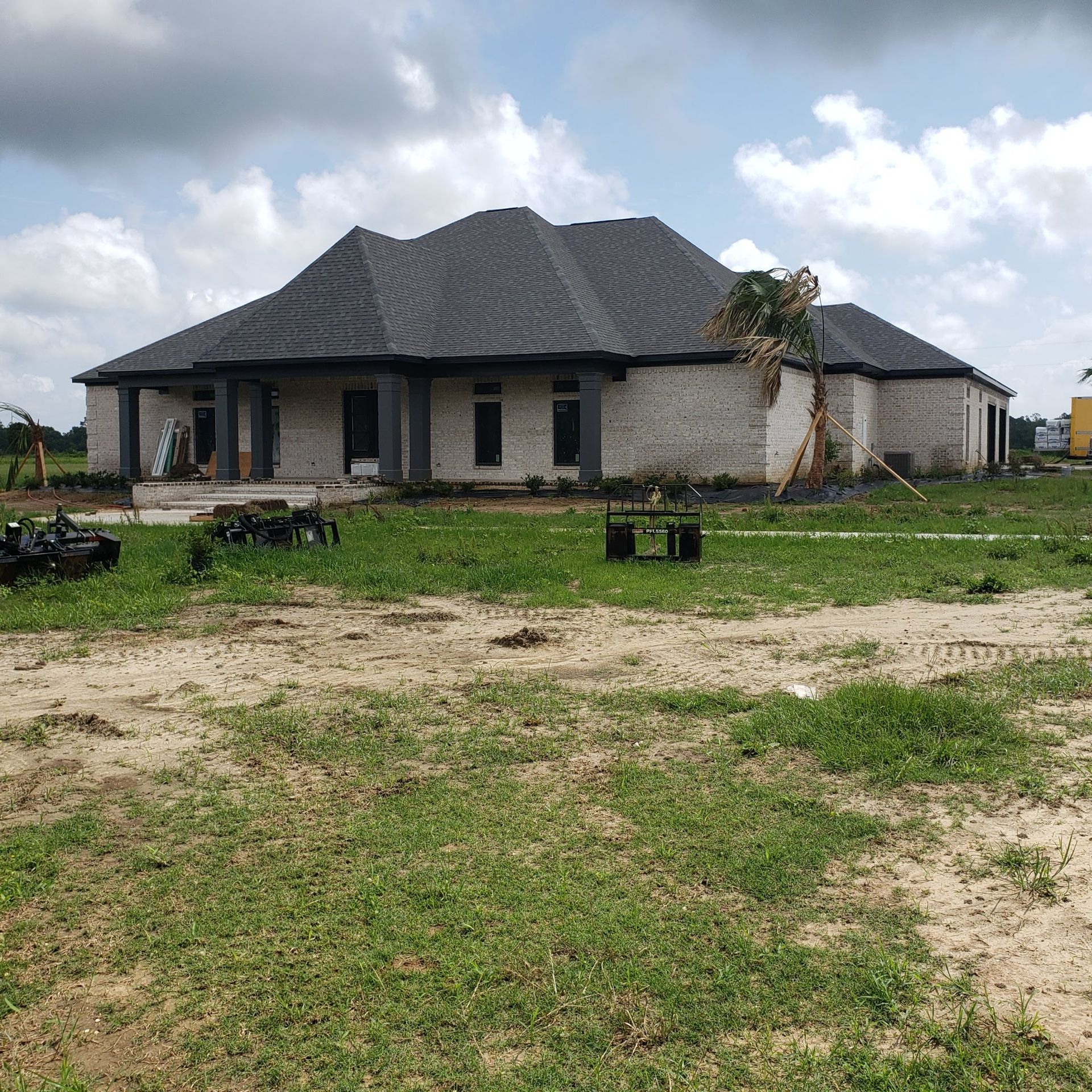 A single-story, light-colored brick house with a dark gabled roof stands on a grassy lot under a cloudy sky.