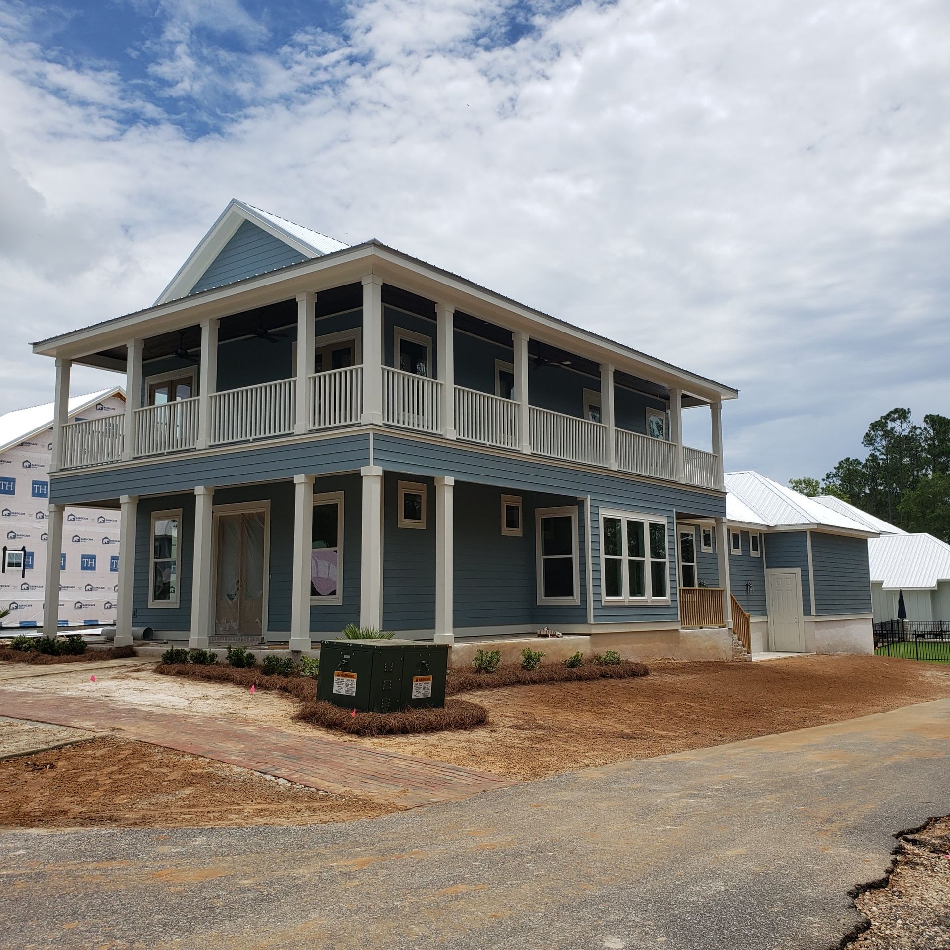 A two-story blue house with white trim and a wrap-around porch under construction, set in a yard with dirt landscaping.