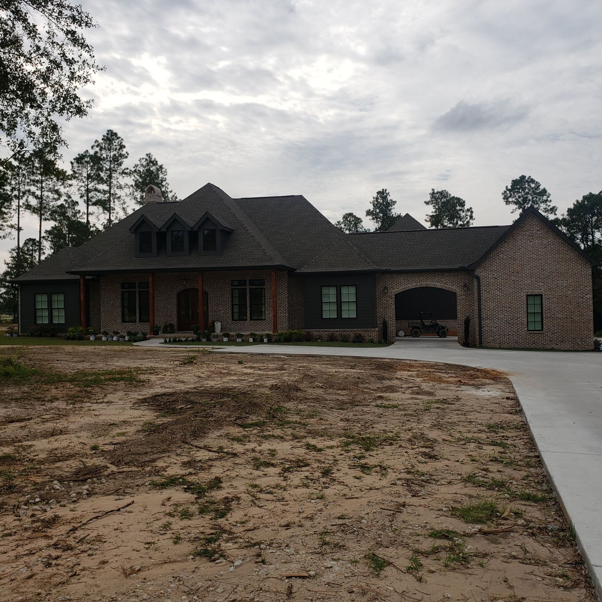 A large, dark-toned luxury home with a stone-accented garage sits on a sandy, undeveloped lot under a cloudy sky.