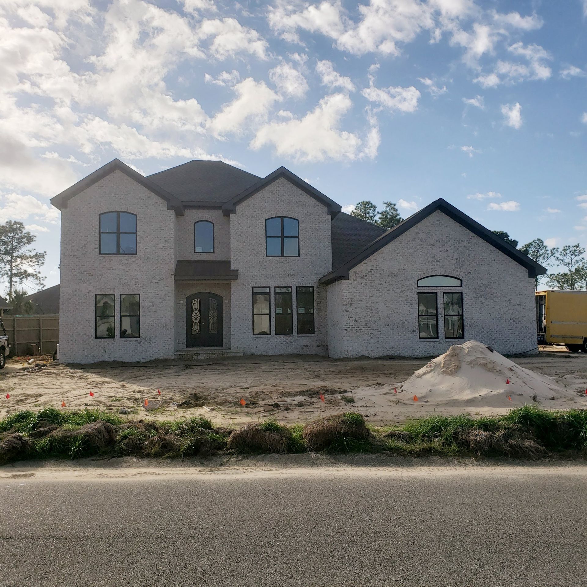 A large, two-story white brick house under construction with a dark shingled roof, set on a sandy lot against a blue sky.