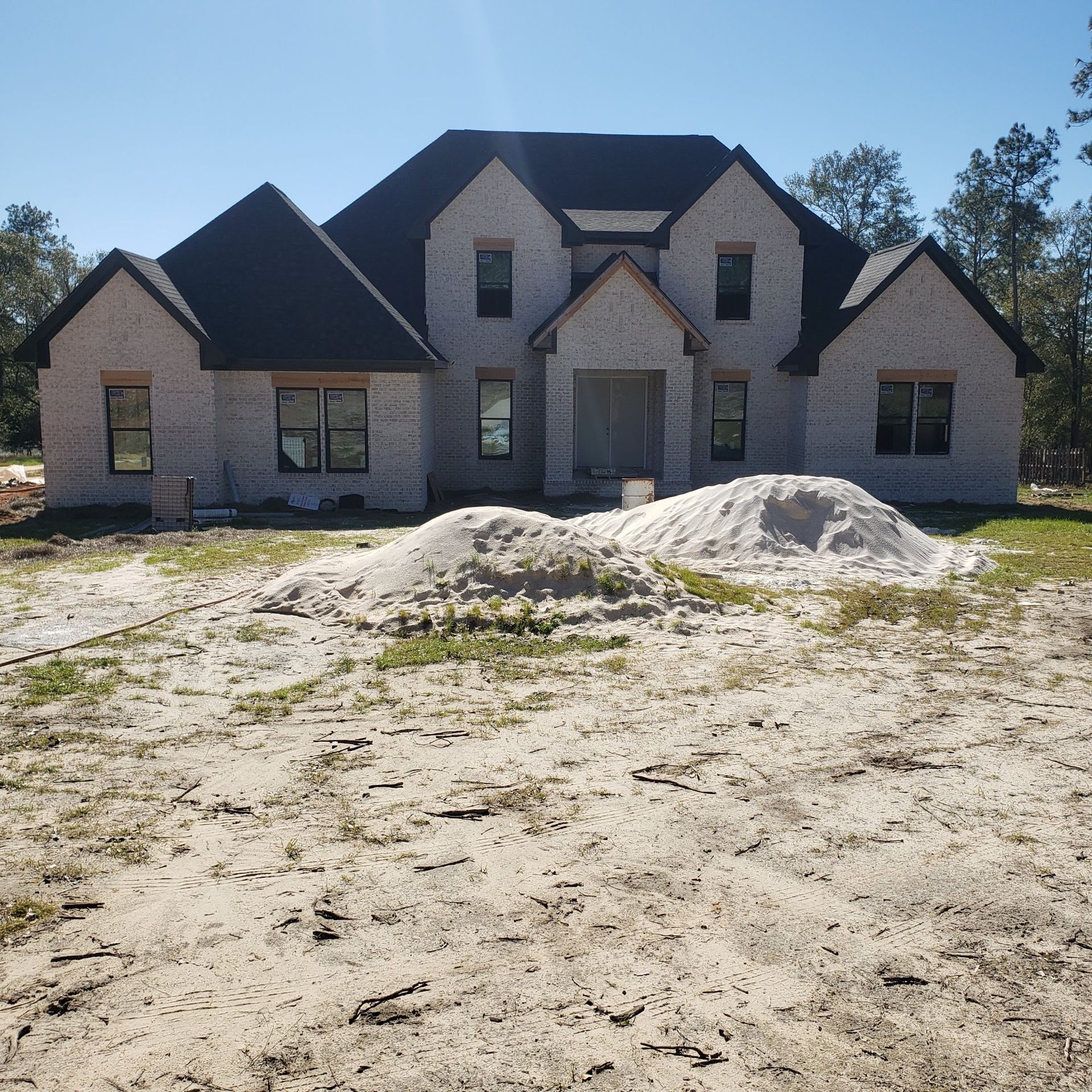 A newly constructed two-story brick house with a dark roof, set against a backdrop of trees, with piles of sand in front.