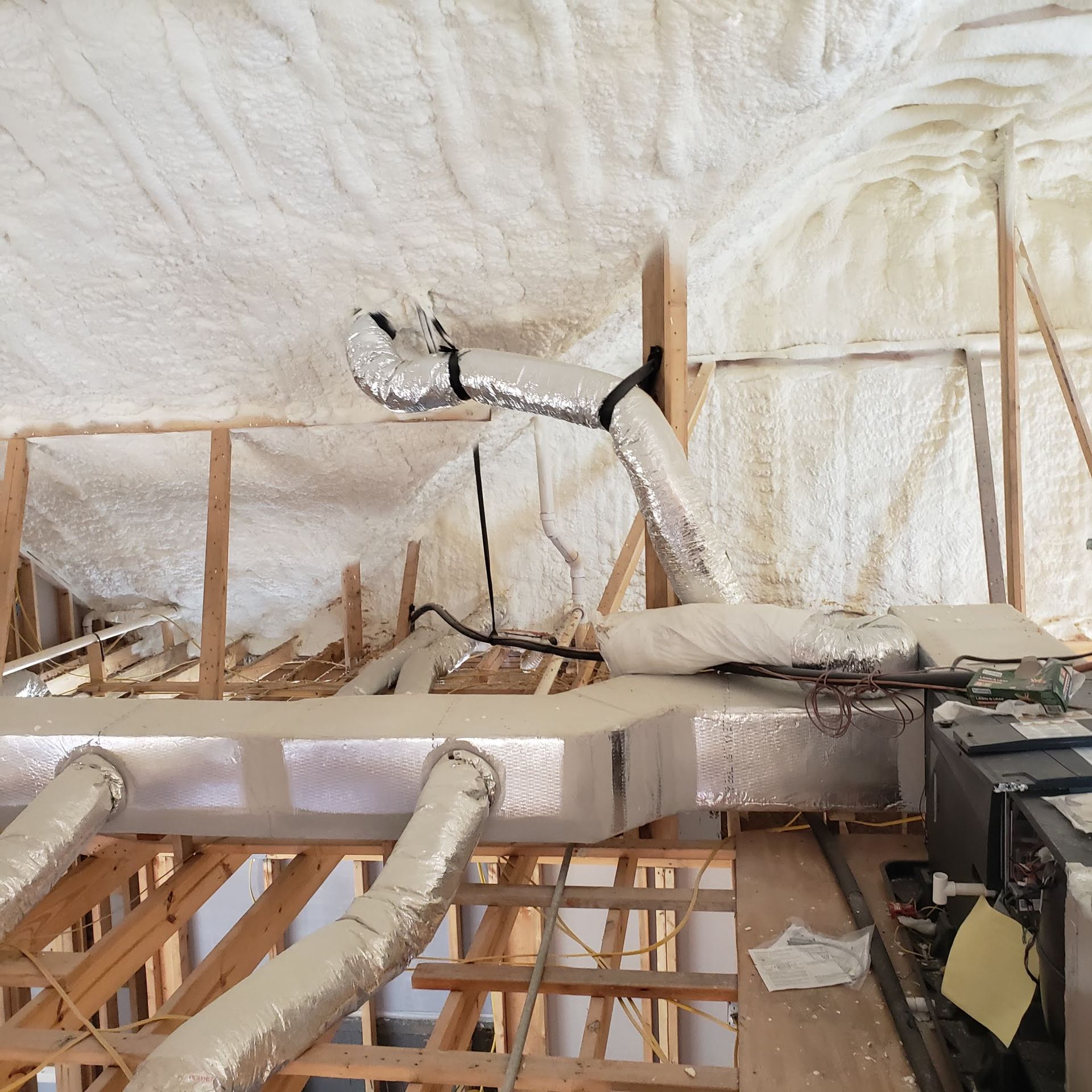 An HVAC unit and insulated air ducts installed in an unfinished attic with spray foam insulation on the roof decking.