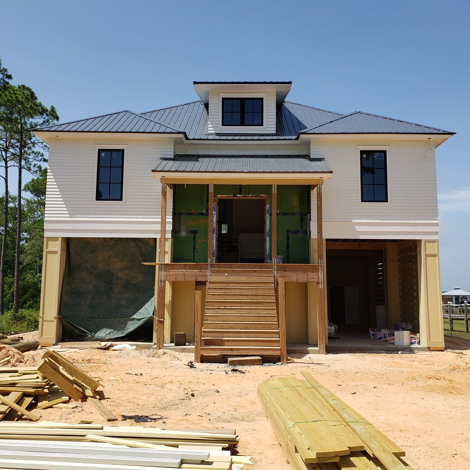 A two-story house under construction with white siding, a metal roof, and an elevated front porch with wooden stairs.