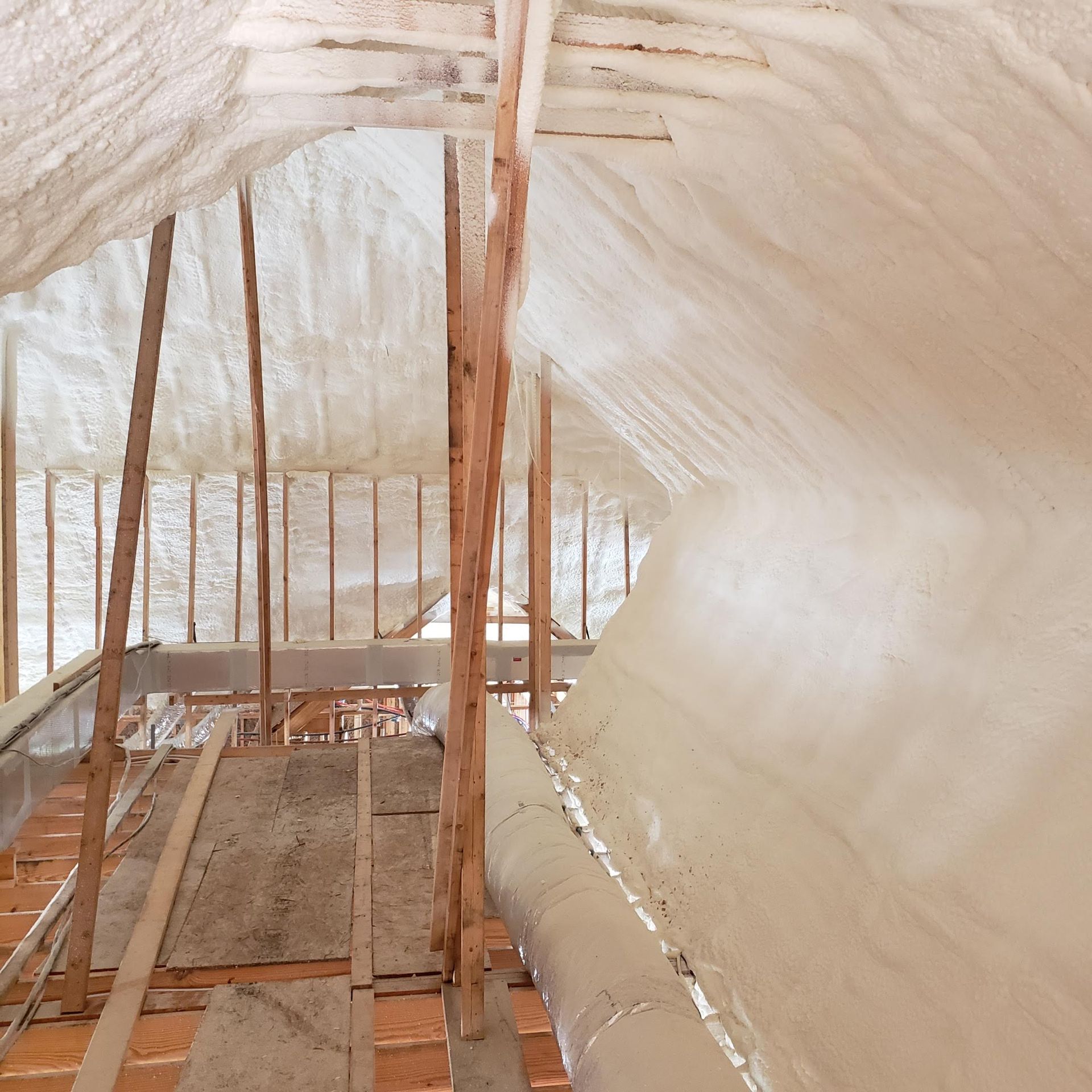 An attic interior features unfinished wooden support beams and flooring covered with thick, sprayed-on white insulation.