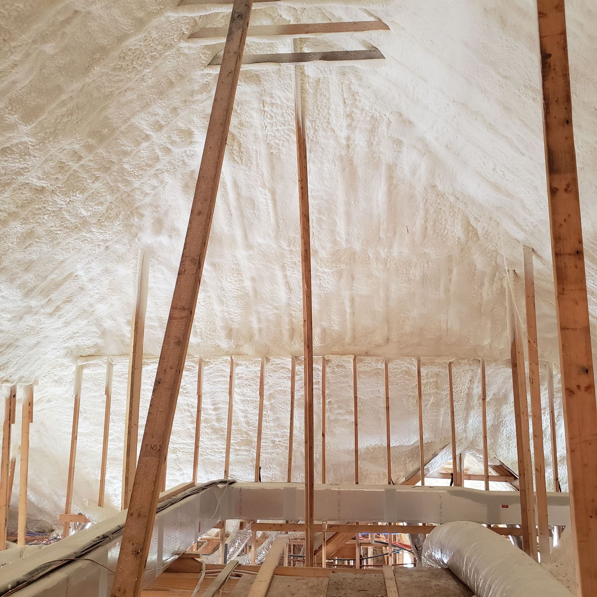 An attic roof structure insulated with white spray foam, featuring exposed wooden support beams and rafters.