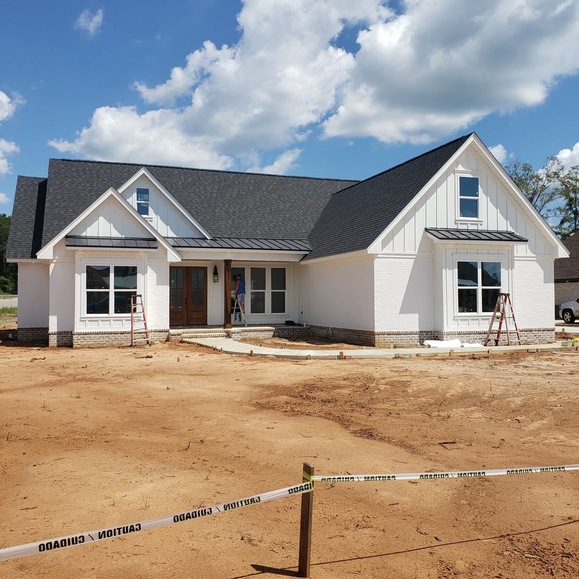 A newly constructed, single-story white house with a dark gray shingled roof, set on a dirt lot under a bright blue sky.