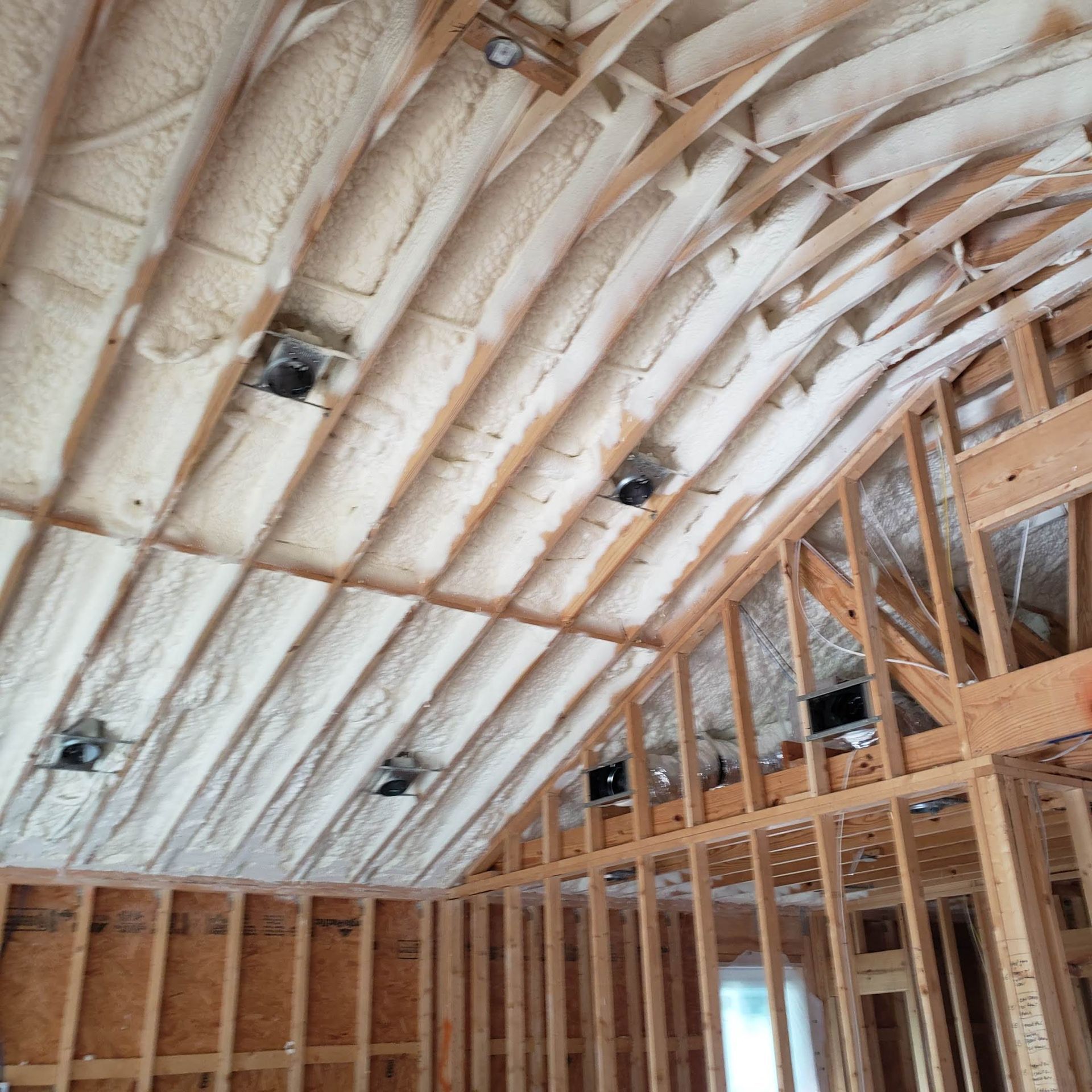 Construction site interior showing exposed wooden wall framing, sloped ceiling beams, and cream-colored spray foam insulation.
