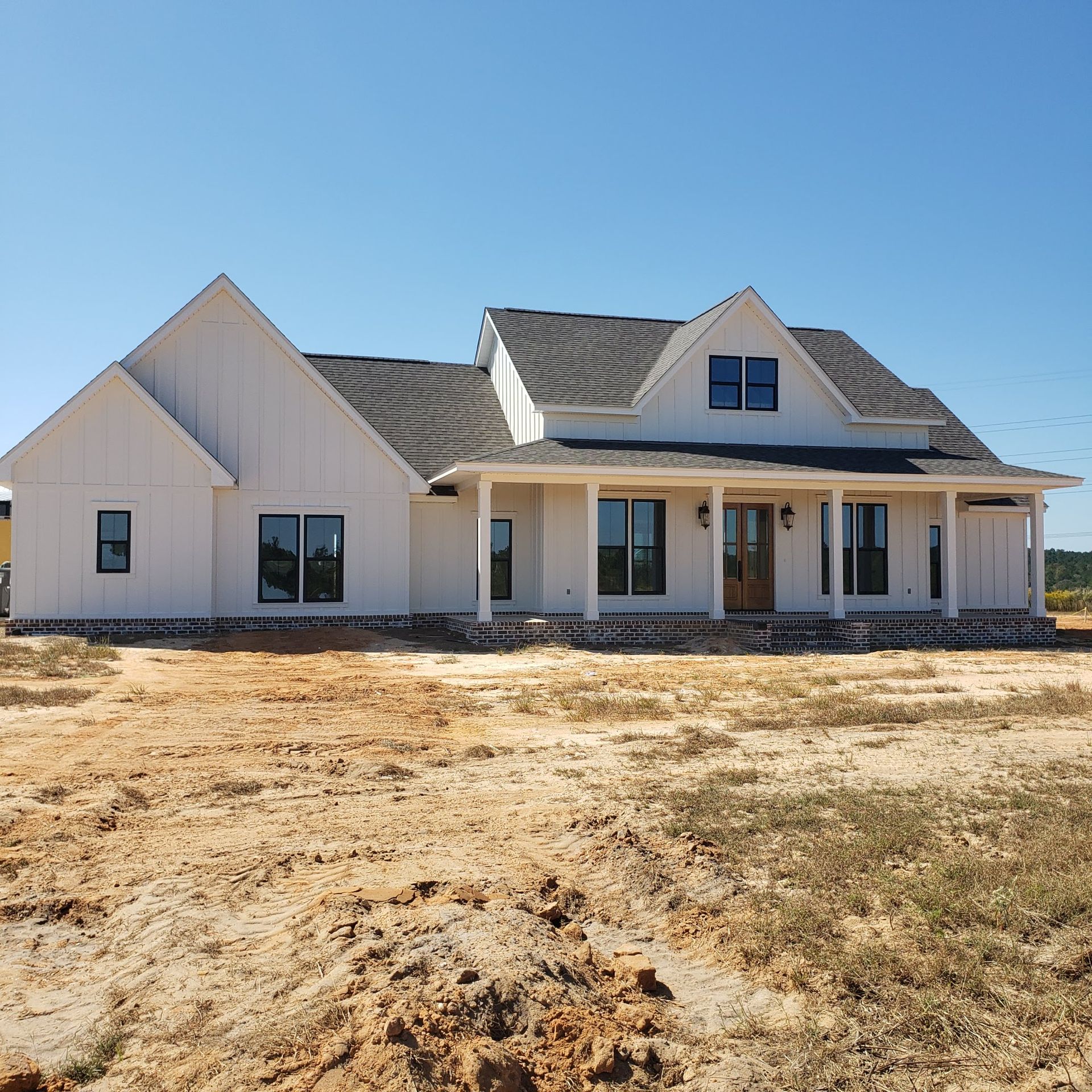 A newly built, one-story white farmhouse with a dark roof and a front porch, situated on a dirt lot under a blue sky.