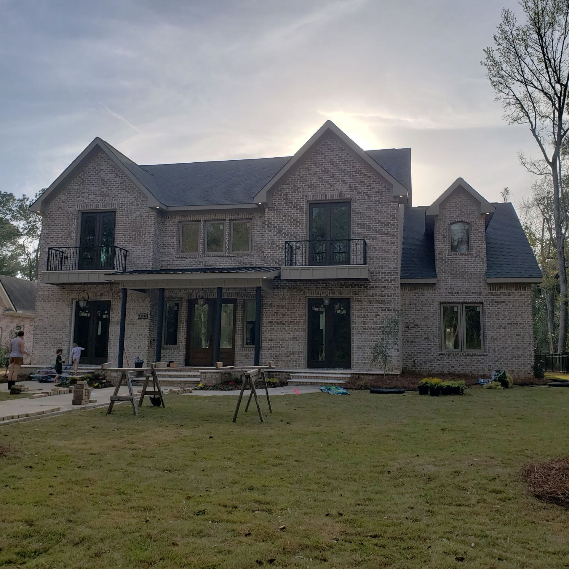 Two-story brick house with a covered patio, balconies, and black window frames under a bright, partly cloudy sky.