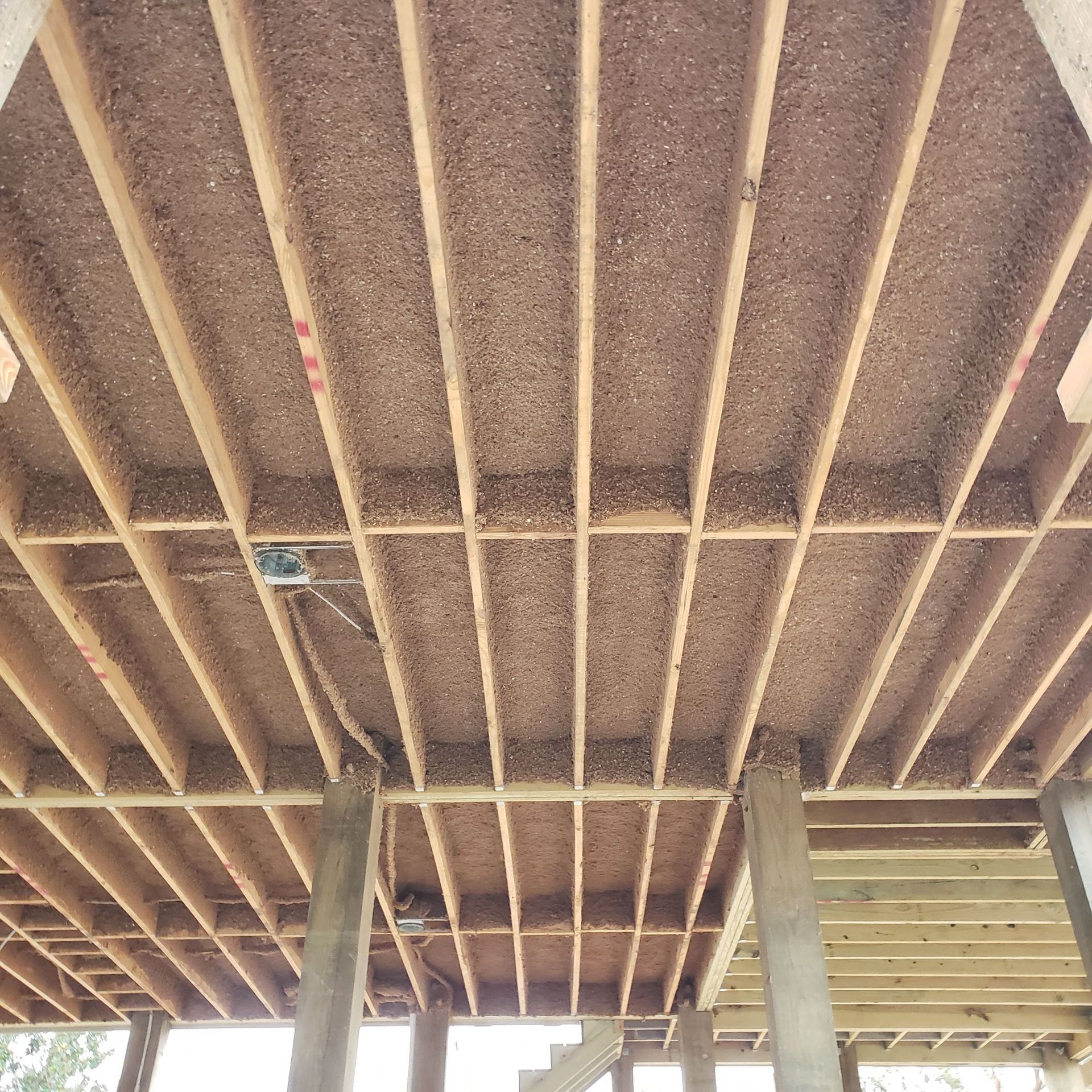 A view from below of wooden deck joists, support posts, and the underside of subflooring materials.
