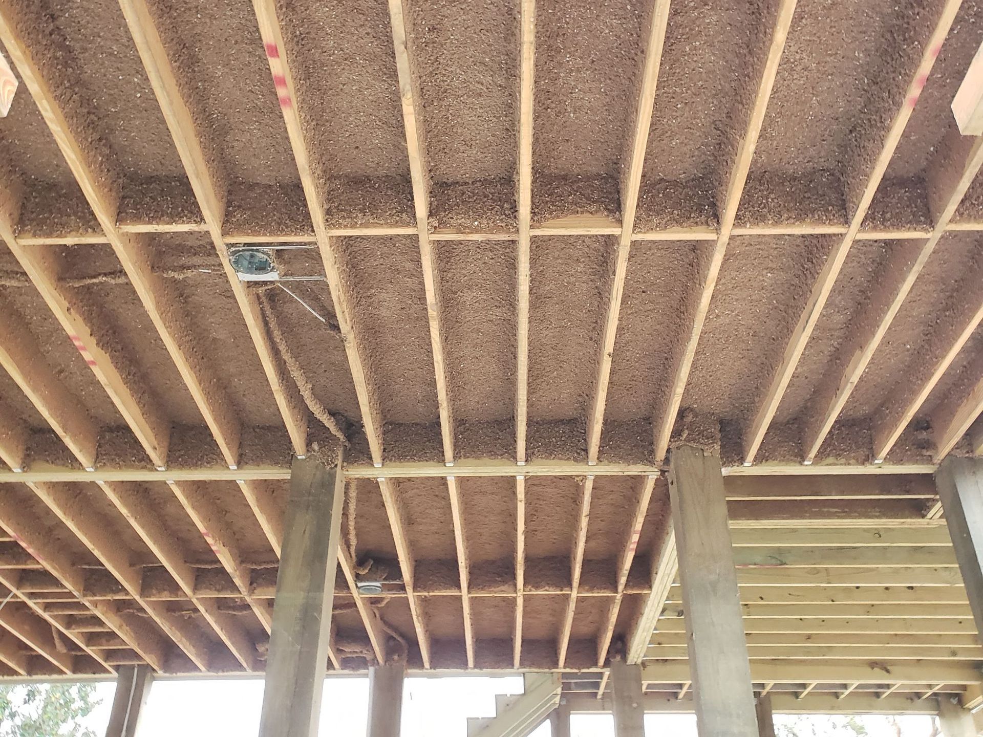 A low-angle view of wooden floor joists beneath an elevated deck, showing framing, structural supports, and wiring.