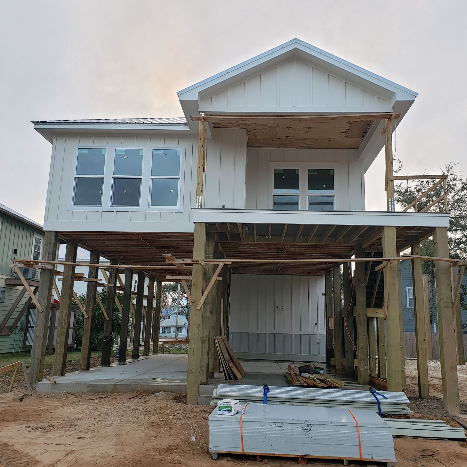 A partially constructed white house on stilts at a sandy building site, with stacks of construction materials in front.