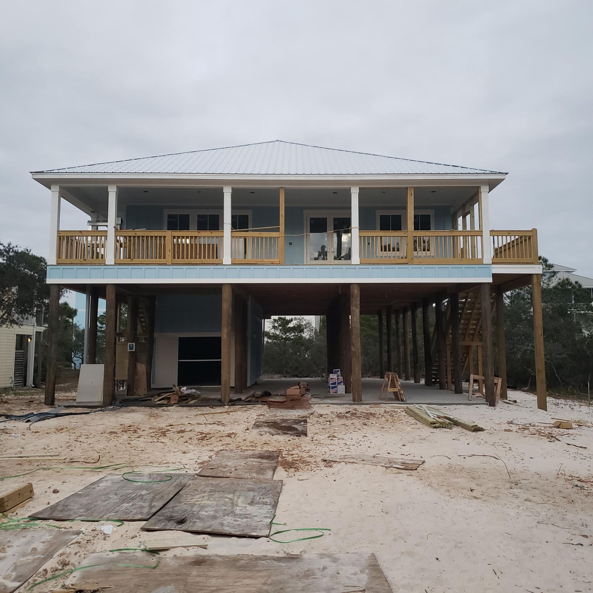 A light blue house on stilts with a wooden deck and white metal roof stands on a sandy lot under a cloudy sky.