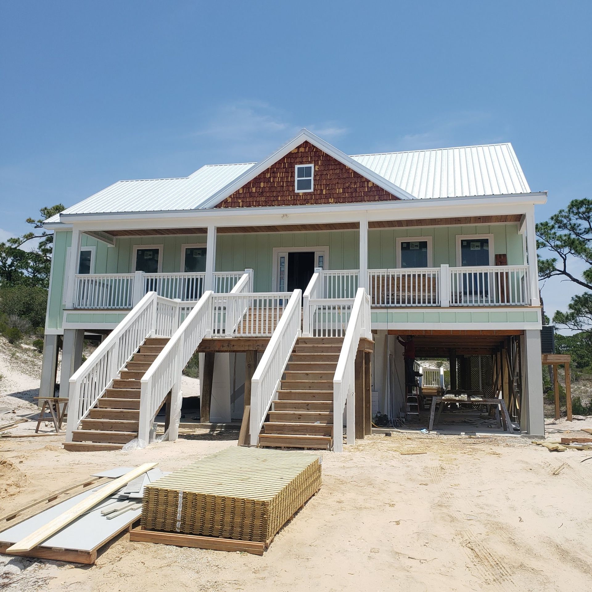 A light green elevated house with a white roof and two symmetrical front staircases sits on a sandy lot under a blue sky.