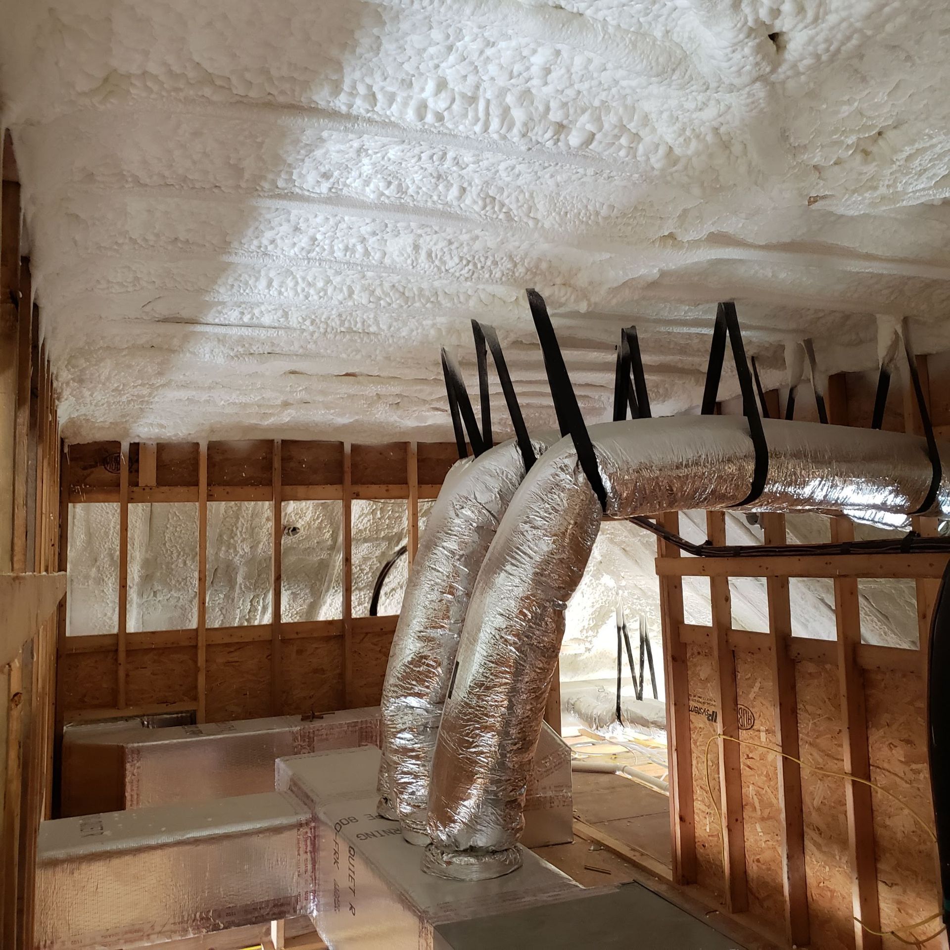Flexible metal HVAC ducts suspended from a ceiling insulated with white spray foam in a wooden attic frame.