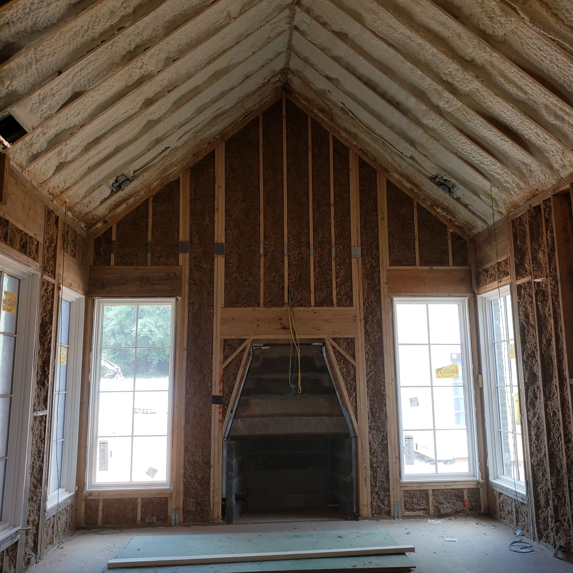 A room under construction with wood framing, spray foam insulation, a central fireplace, and two tall windows.
