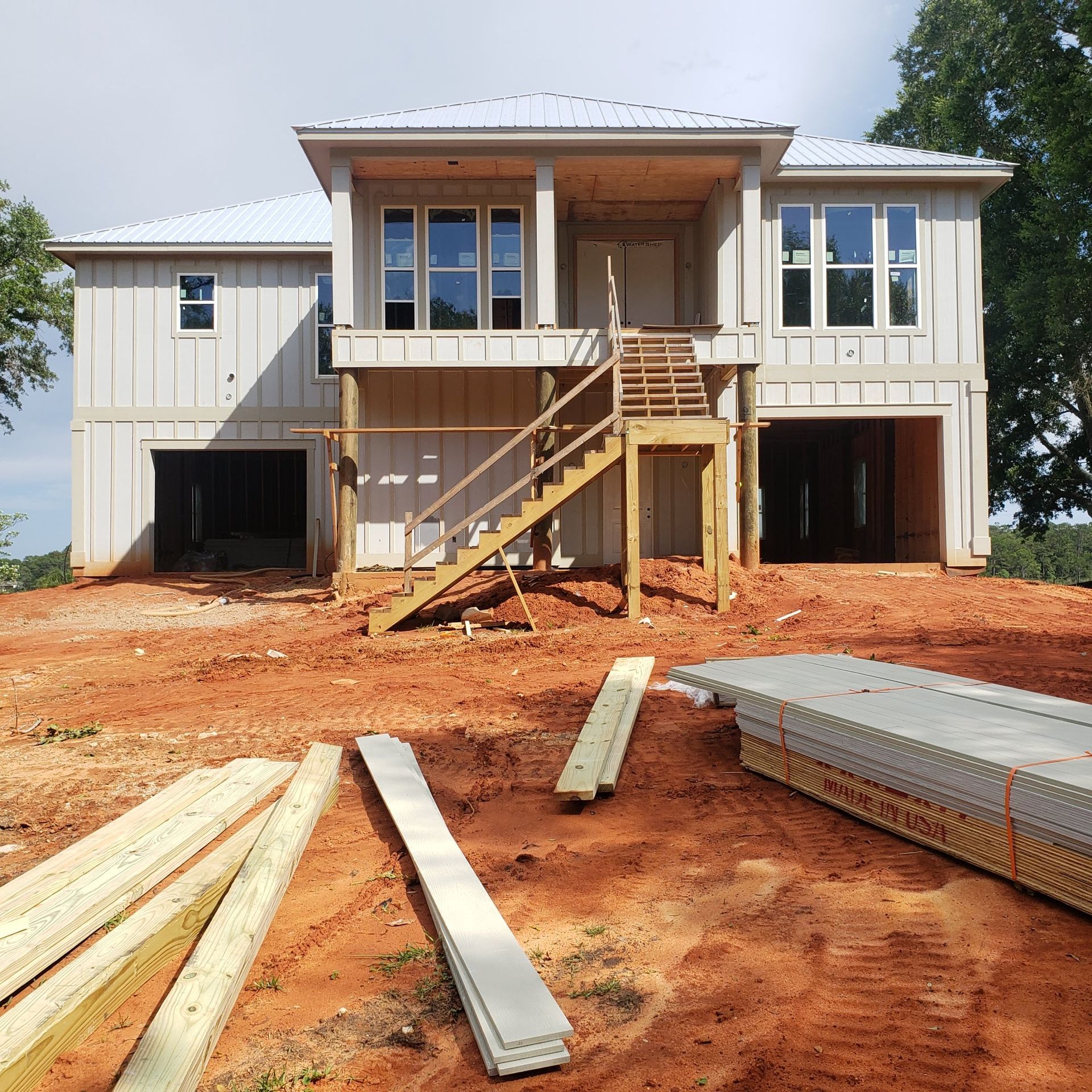 A two-story house under construction with light gray vertical siding, a white metal roof, and wooden stairs in front.