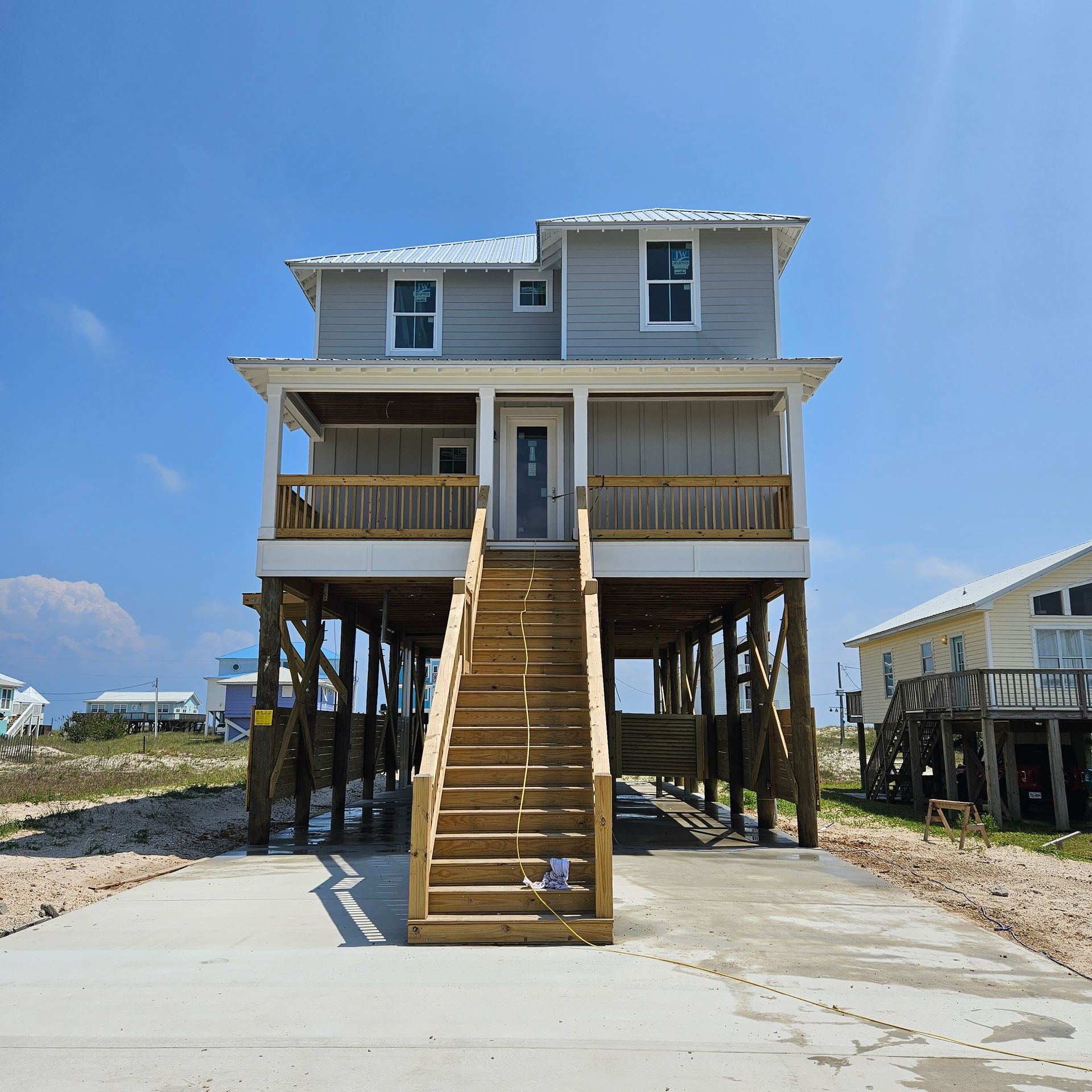 A two-story grey elevated house on stilts with a central wooden staircase, situated on a concrete lot under a clear sky.